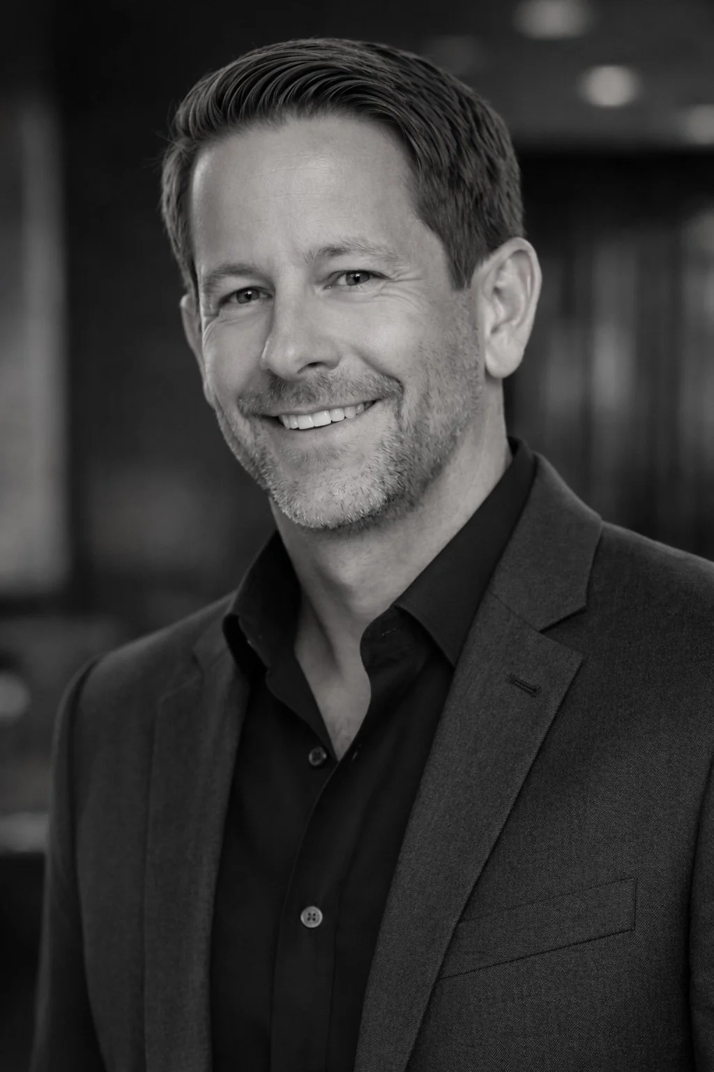 Black and white professional portrait of a smiling man with short hair and beard, wearing a dark suit and shirt, in an indoor setting