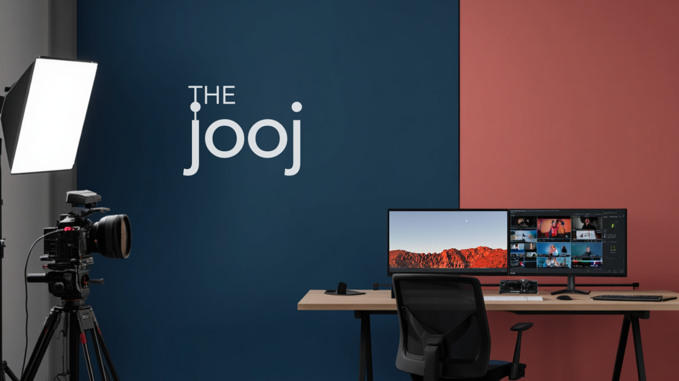 A video production studio setup with a camera on a tripod, softbox light, two computer monitors displaying video editing screens, a desk with a keyboard, mouse, and a small camera, and a blue and red wall with "The Jooj" logo.