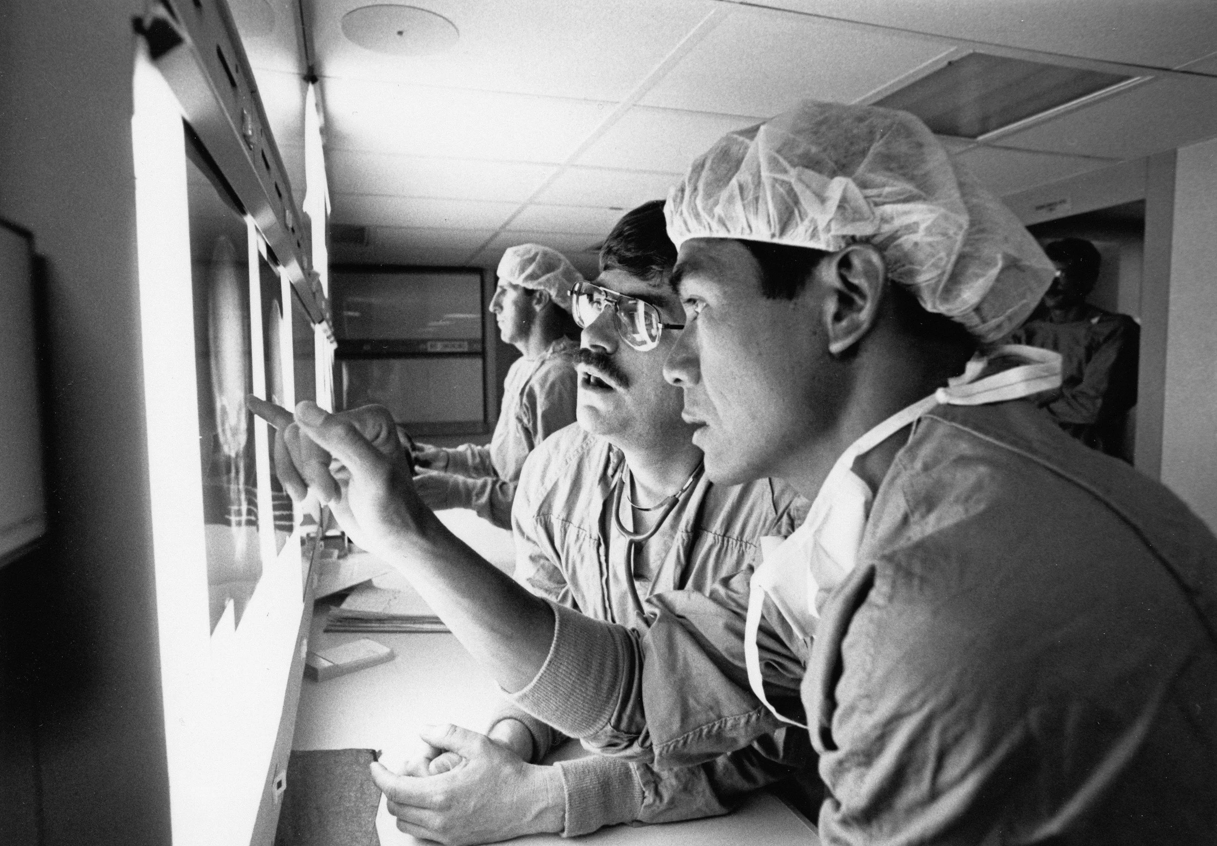 Two medical professionals, a man and a woman, wearing protective hair covers, scrubs, and glasses, are working with a computer or medical device in a clinical setting.