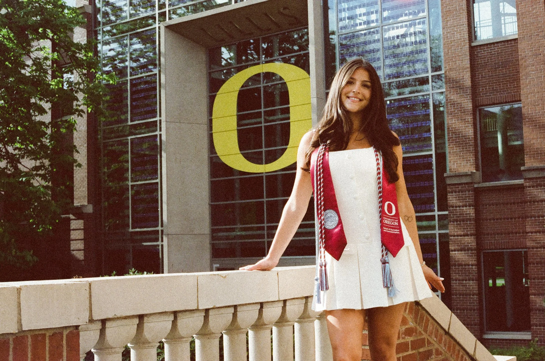 A young woman in a white dress celebrating graduation, wearing a red and white honor stole and cords, standing on a balcony with a university building and large yellow letter O in the background.