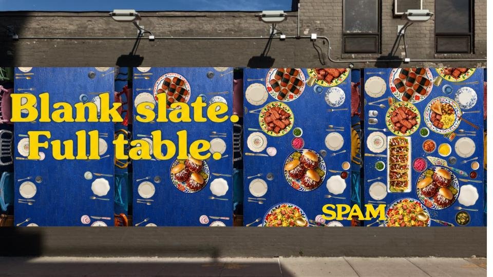 Colorful outdoor art display with painted tables and plates showing various foods; bold yellow text reads "Blank slate. Full table." and "SPAM."