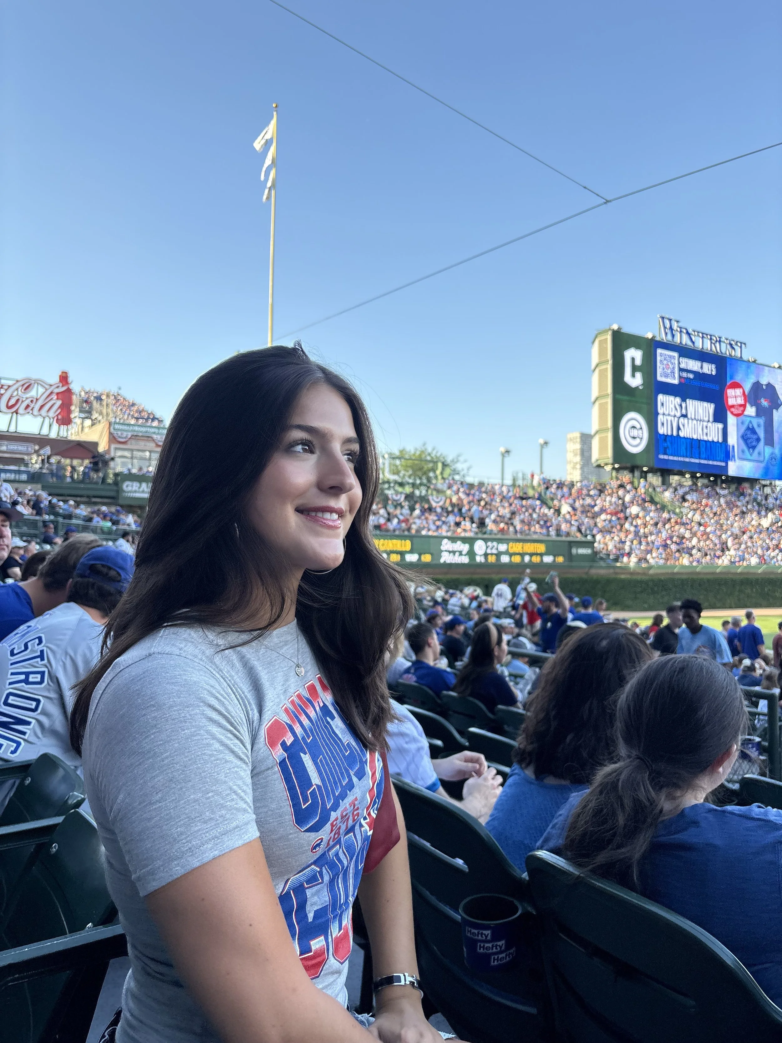 A young woman sitting in a stadium during a baseball game, wearing a gray T-shirt with red, white, and blue text, and smiling while looking towards the field.