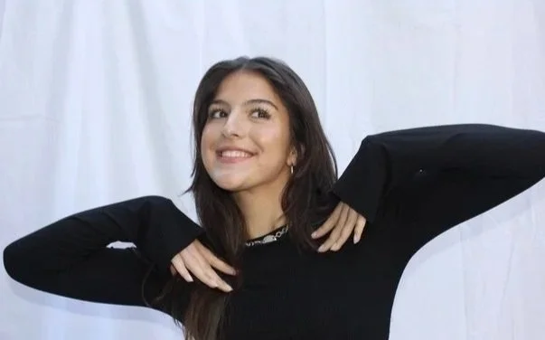 A young woman with long brown hair smiling and posing with her hands near her face, wearing a black long-sleeve top against a plain white background.