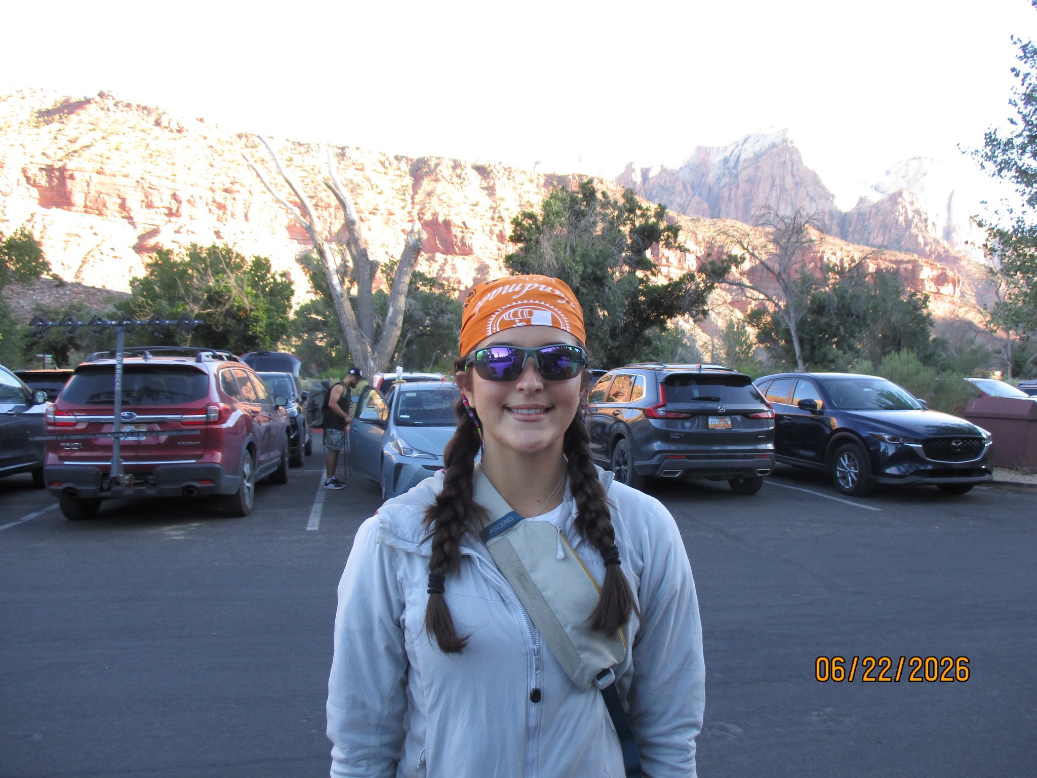 A woman with long braided hair, wearing sunglasses, a light gray jacket, and an orange bandana, standing in a parking lot with red rock cliffs in the background. The date 06/22/2026 is displayed in the bottom right corner.