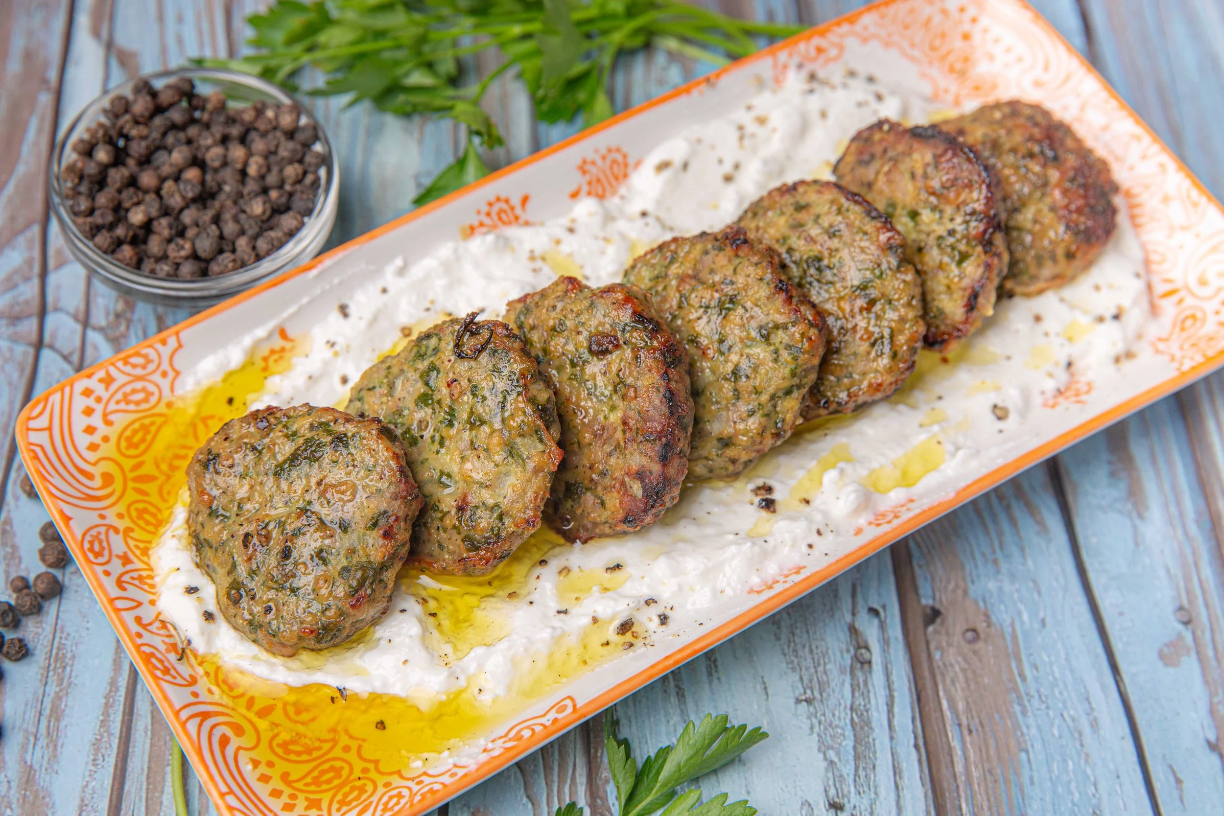 Plate of grilled meatballs with herbs drizzled with olive oil, served in a decorative orange and white dish, on a rustic wooden table with fresh parsley and black peppercorns.