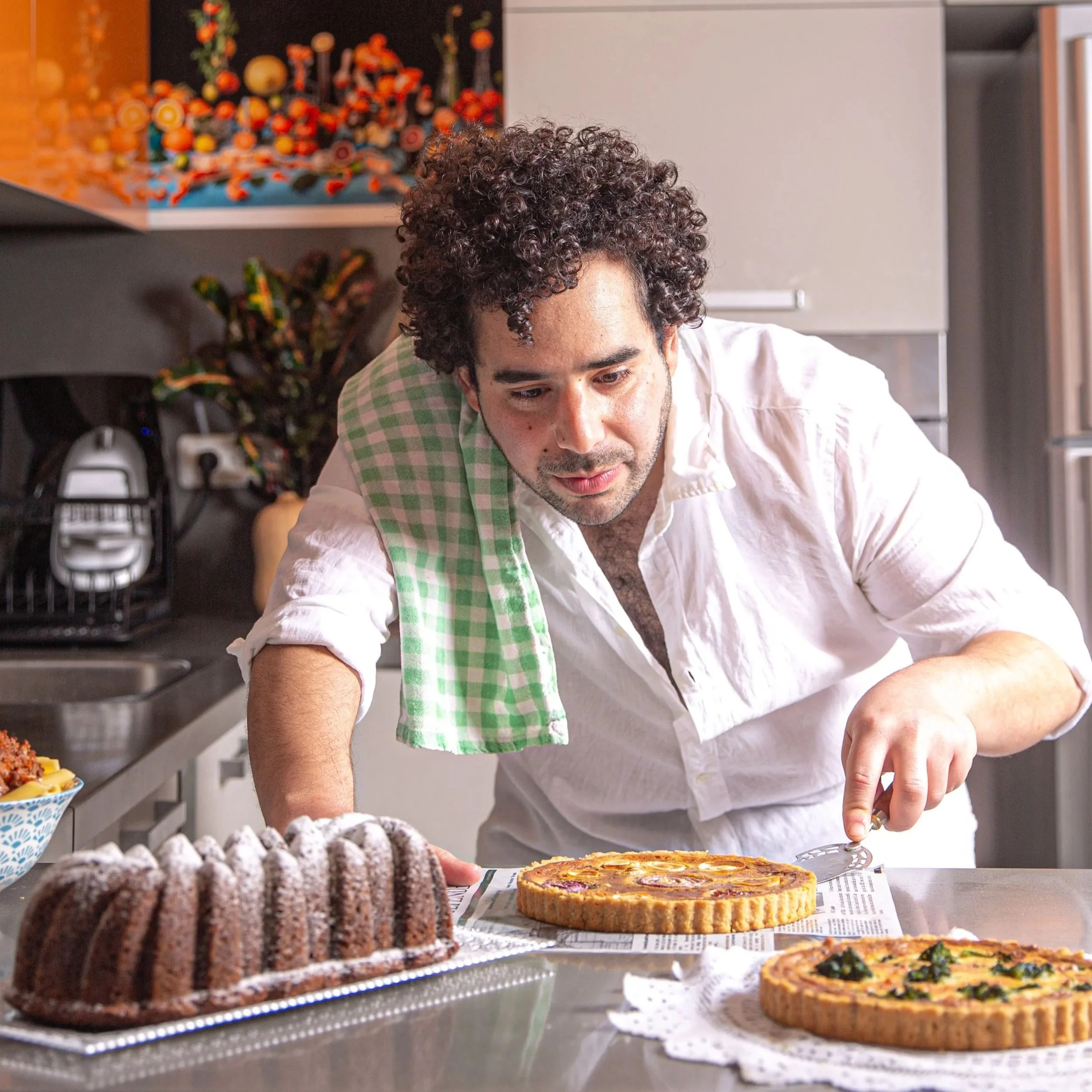 Chef Gal Ben Moyal stands behind a table of desserts, including cakes and pastries, in a kitchen.