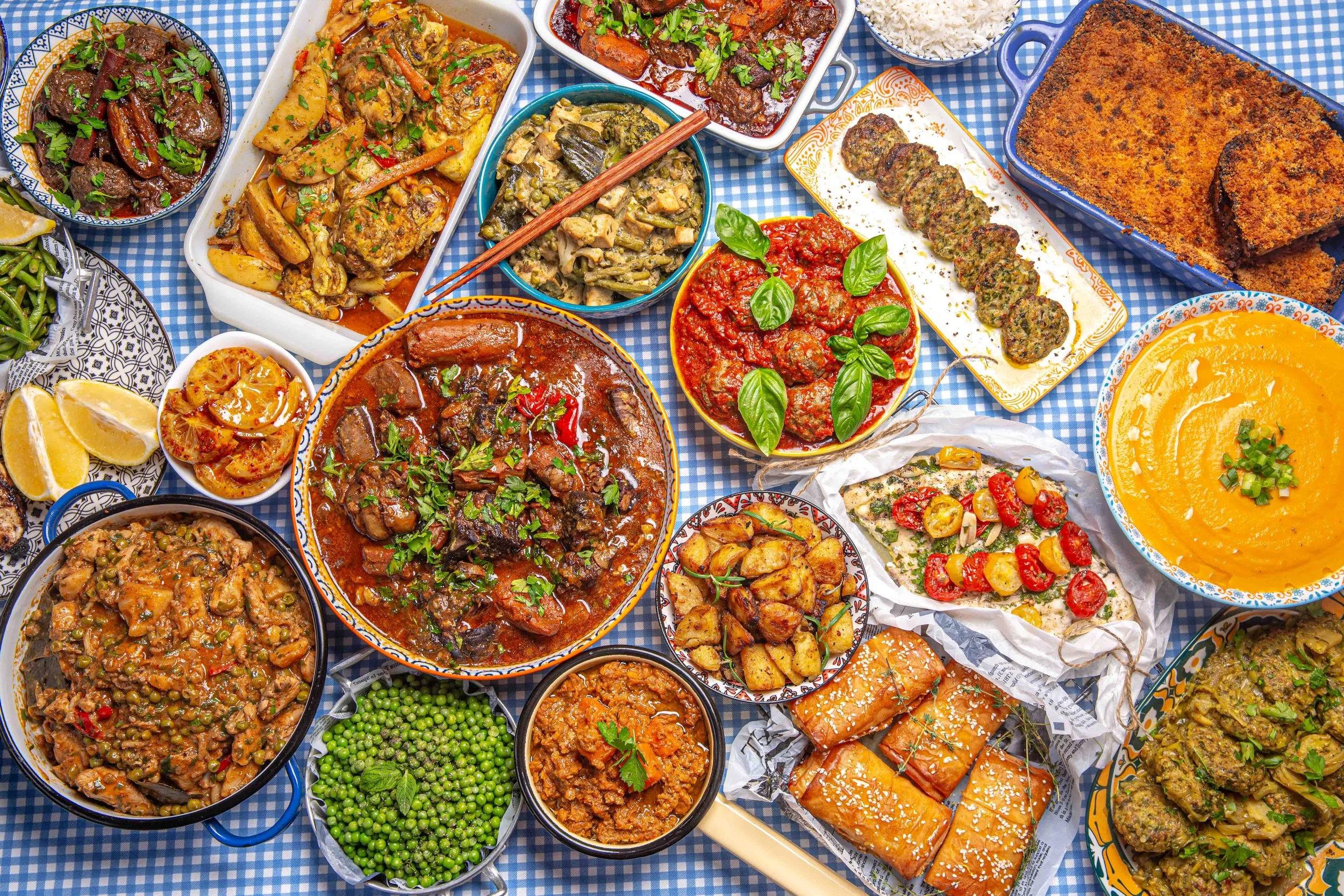 A variety of Mediterranean dishes served in bowls and on plates, including fish, vegetables, rice, and bread, placed on a blue checkered tablecloth.