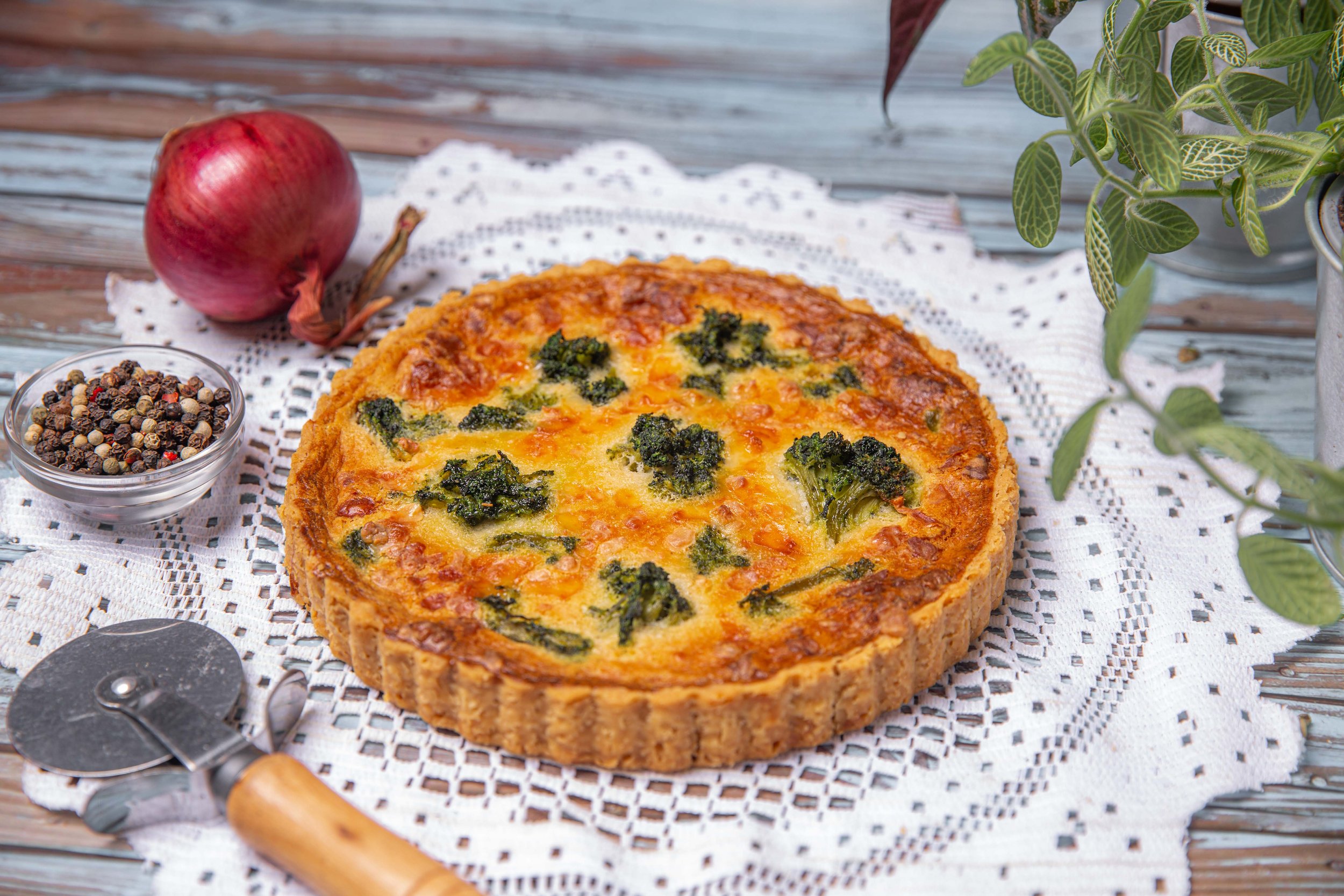 A freshly baked quiche with broccoli on a decorative white lace placemat, surrounded by a red onion, a small bowl of mixed peppercorns, with potted greenery in the background, on a wooden surface.