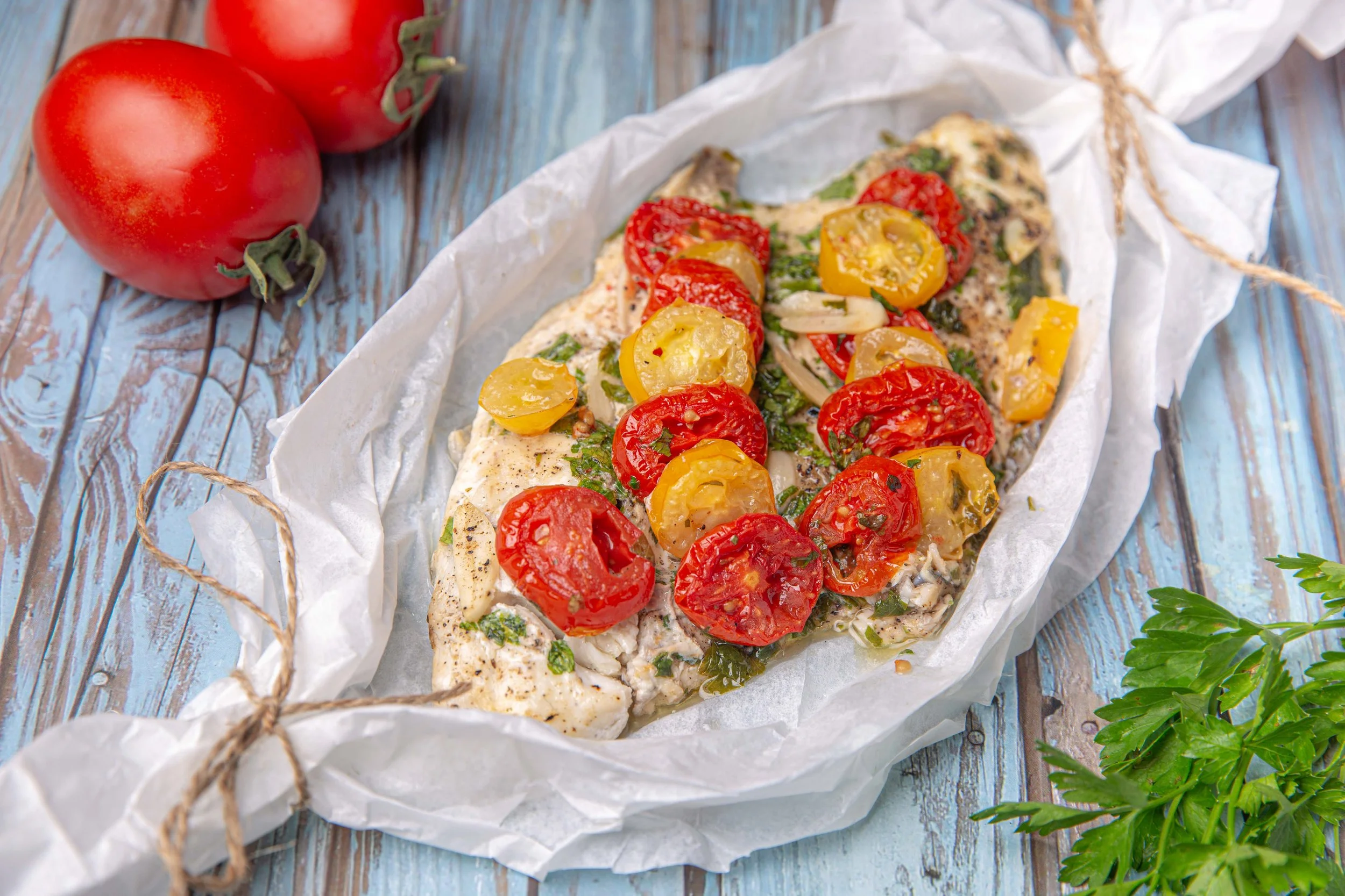 Baked fish fillet topped with cherry tomatoes, chopped herbs, and sliced onions, wrapped in parchment paper, on a rustic wooden surface with fresh parsley nearby.