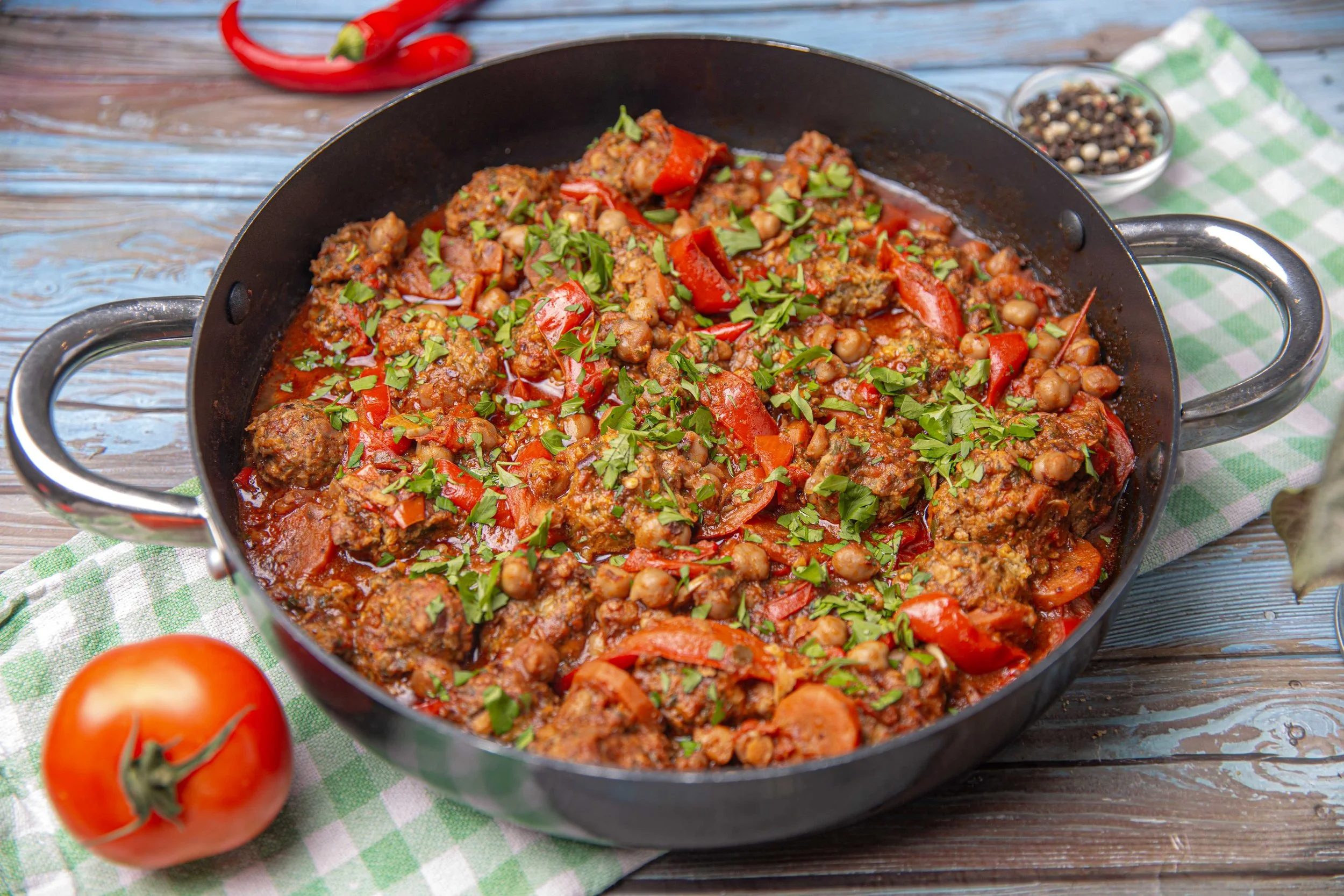 A black skillet filled with a cooked stew containing meat, chickpeas, red bell peppers, and chopped green herbs, set on a rustic wooden table with a green and white checkered cloth nearby. There are some tomatoes and a bowl of peppercorns in the background.