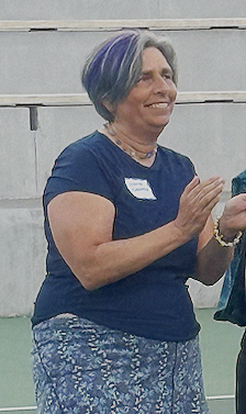A woman with short gray and purple hair wearing a navy blue shirt and patterned pants, smiling and gesturing with her hands outdoors.