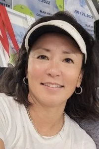 A woman with dark hair, wearing a white cap and earrings, smiling in front of a colorful background at what appears to be a promotional or event setting.