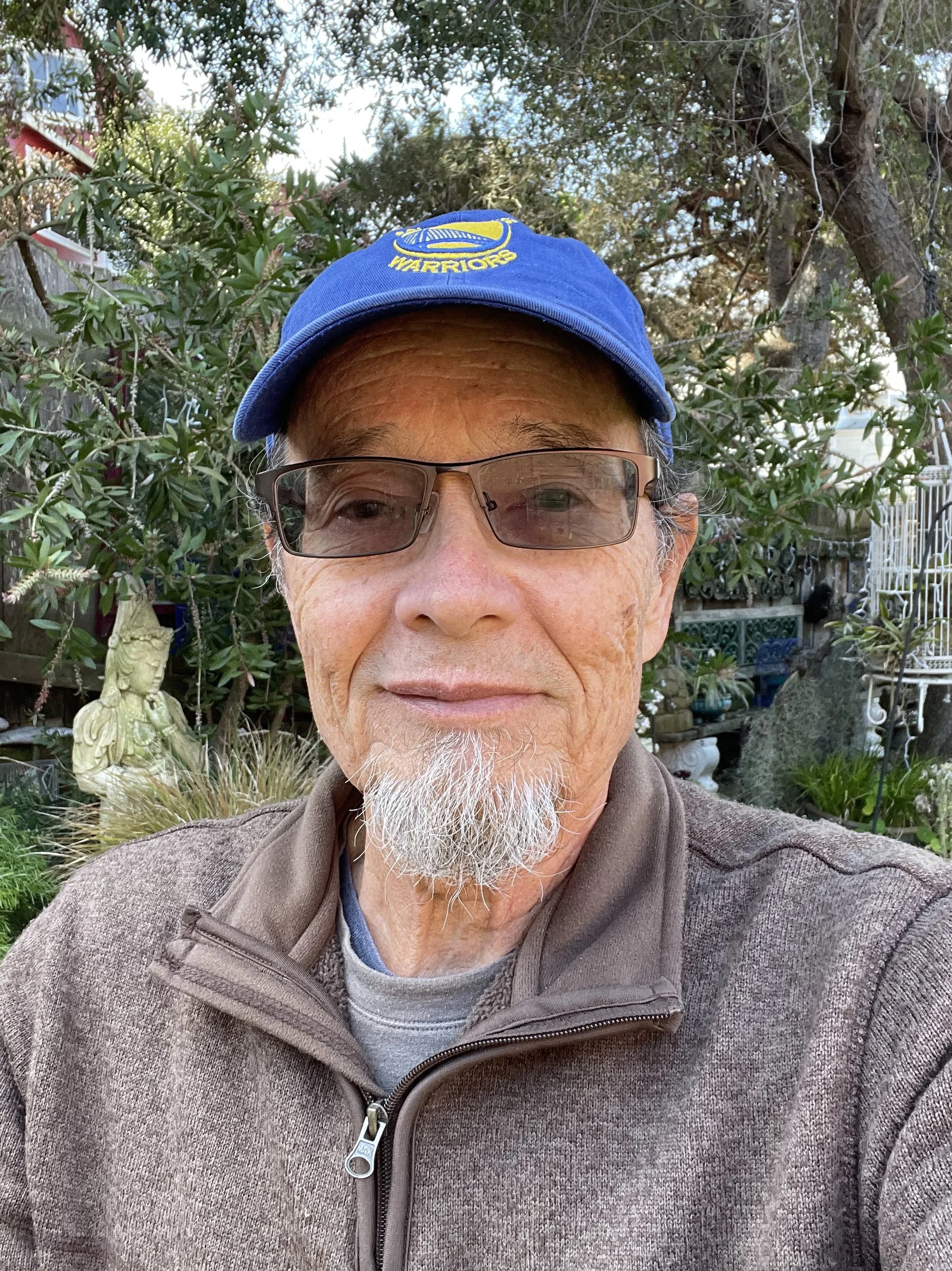 A close-up of a man with glasses and a beard wearing a blue cap with a yellow logo, outdoors with trees and garden decorations in the background.