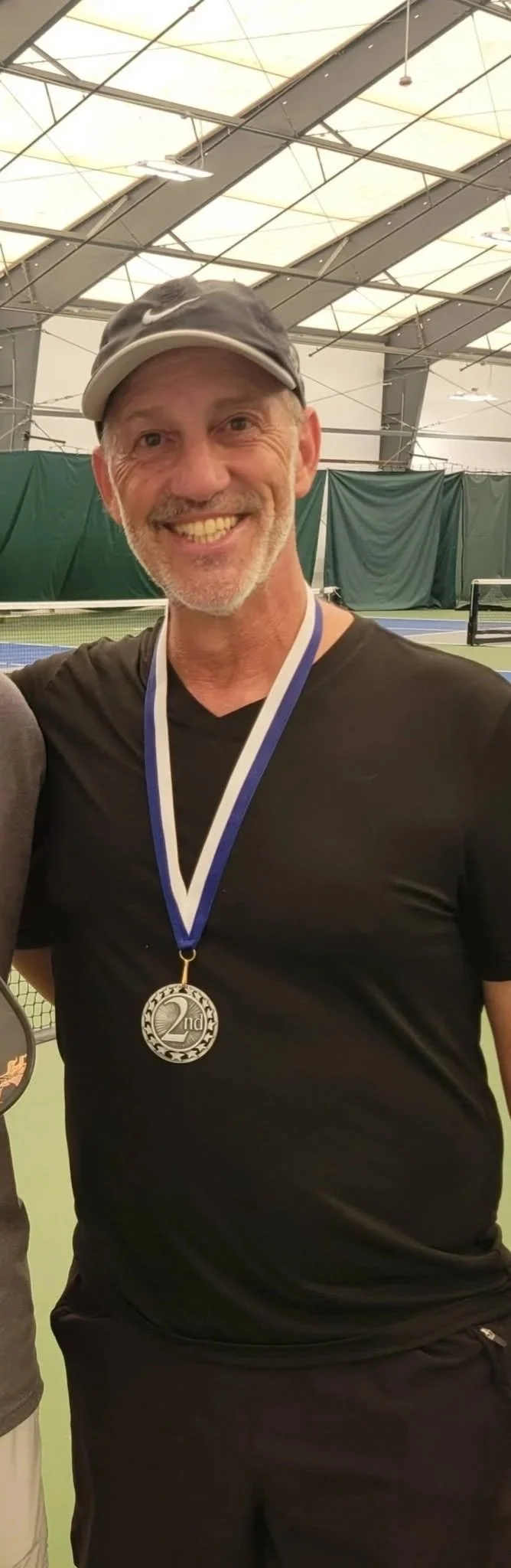 A smiling man wearing a black Nike cap and black athletic shirt, with a silver 2nd place medal around his neck, standing on an indoor tennis court.