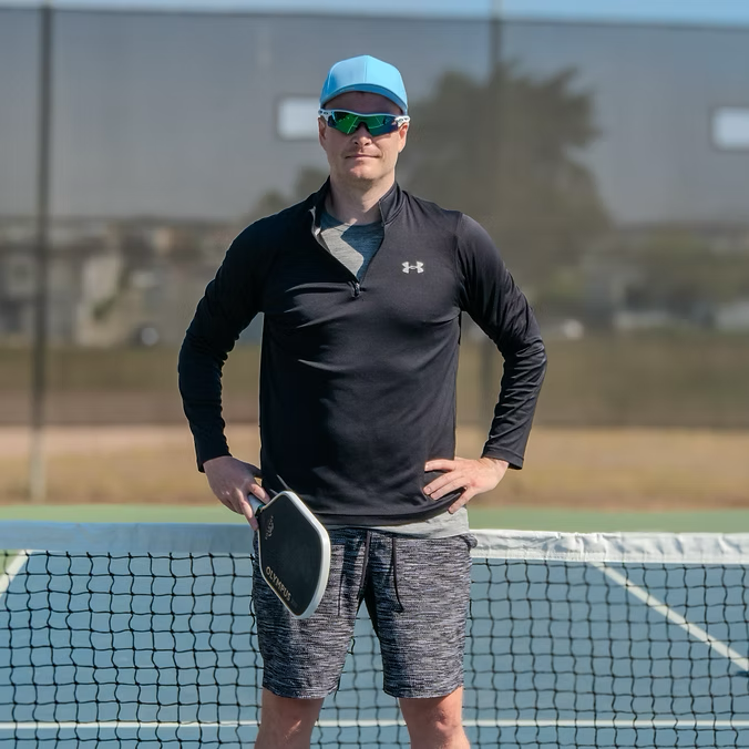 Man standing on tennis court holding ping pong paddle, wearing a light blue cap, sunglasses, black long-sleeve athletic shirt, and gray shorts.