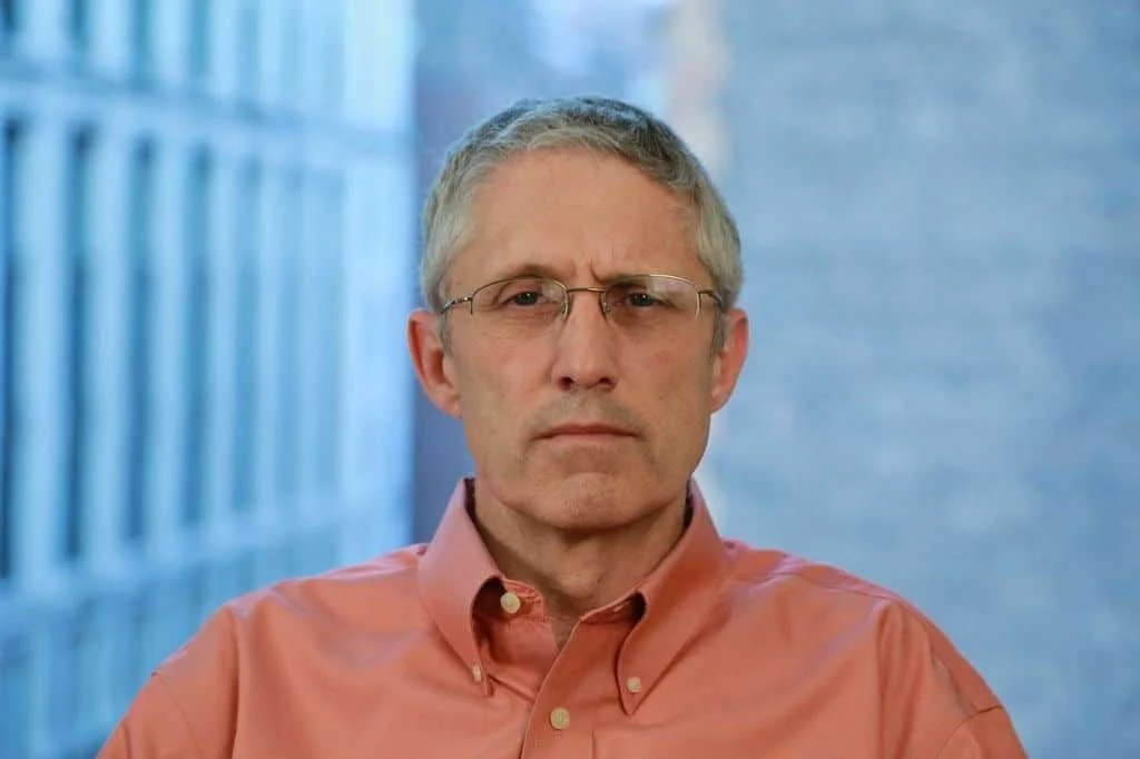 A middle-aged man with gray hair and glasses wearing a salmon-colored button-up shirt, standing in front of a blue background with an out-of-focus architectural structure.