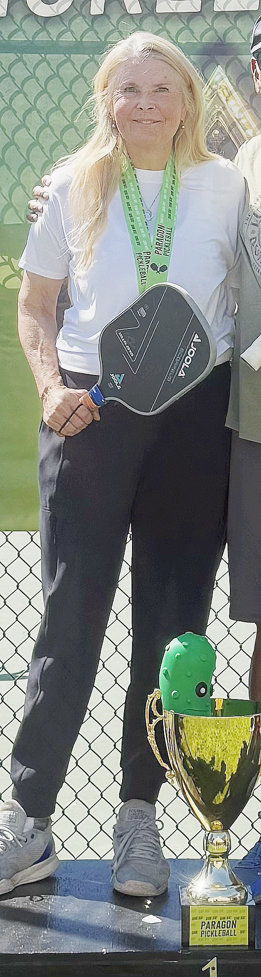 A woman with long blonde hair wearing a white t-shirt, black pants, and gray sneakers, holding a paddle, standing in front of a green fence with a tennis court behind her. She has a green lanyard around her neck and is smiling. There is a green pickle-shaped toy and a gold trophy with a label that reads 'Paragon Pickleball' in front of her.