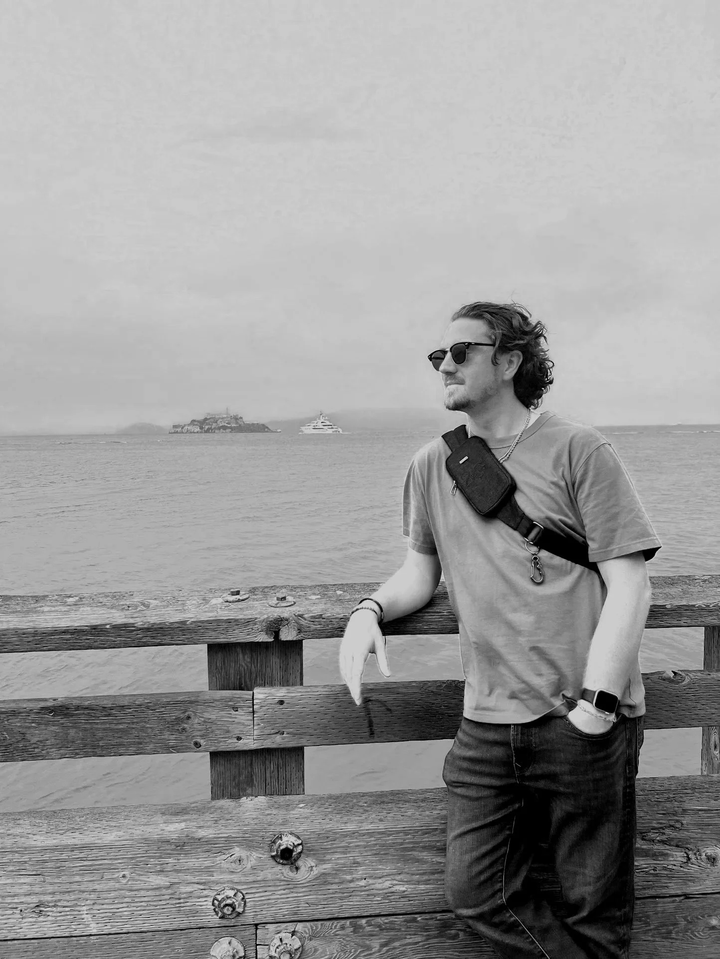 A man in sunglasses leaning on a wooden railing by the water, with islands and a yacht in the background.