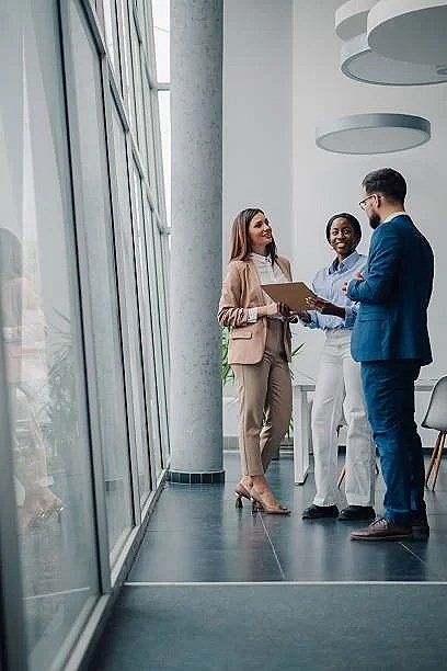 Three business professionals having a discussion in a modern, glass-walled office space with large windows and ceiling decor.