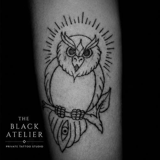 Black and gray tattoo of an owl with horns and detailed eyes, perched on a branch with a leaf and a design resembling an eye on the branch, surrounded by radiating lines.