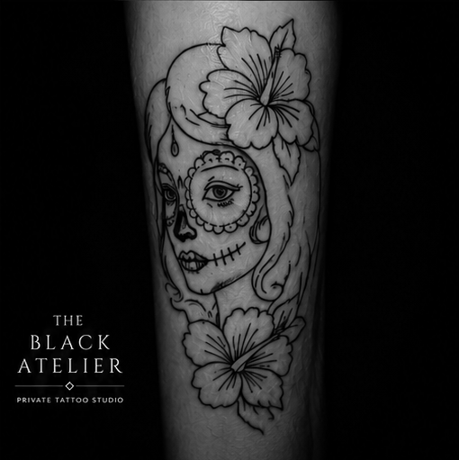 Tattoo of a woman's face with sugar skull makeup, surrounded by hibiscus flowers, on a person's arm.