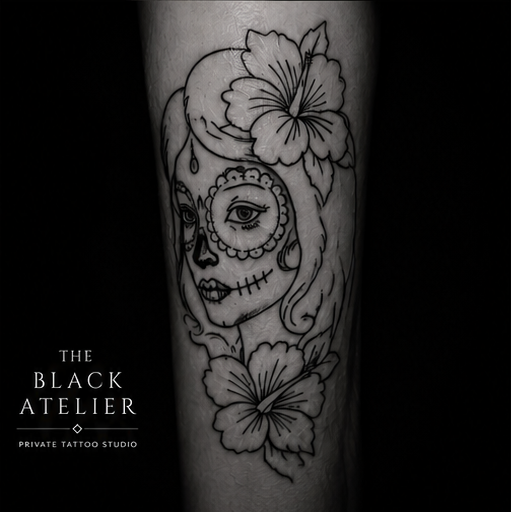 Black and white tattoo of a woman's face with a floral design, resembling a sugar skull or Dia de los Muertos style, on a person's arm.