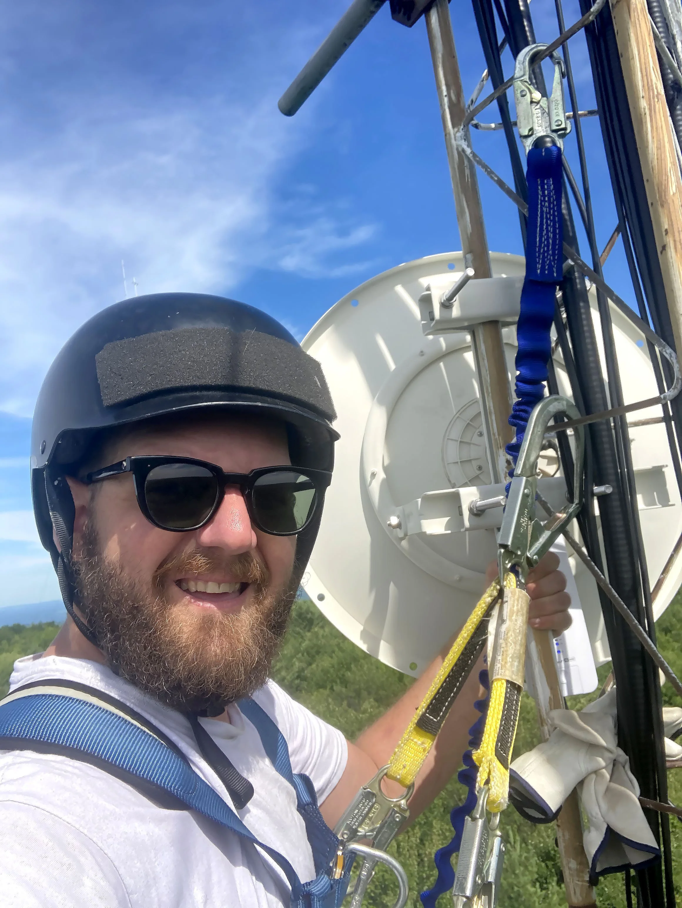 Speiser with a beard and sunglasses wearing a helmet and white shirt, smiling, taking a selfie at the top of a telecommunications tower with blue sky in the background.
