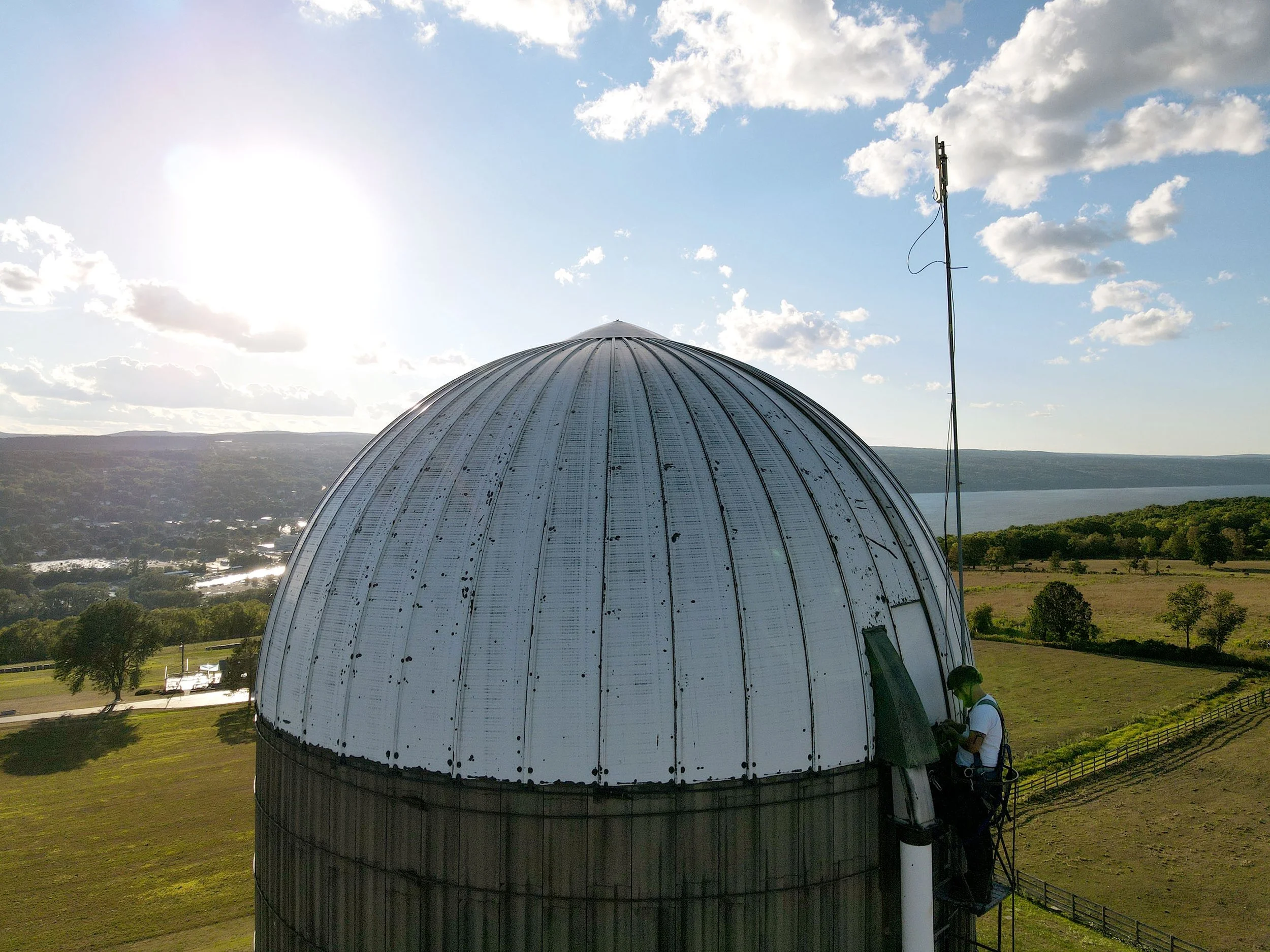 The top of a silo with Speiser next to it, set against a landscape of trees, hills, and Lake Cayuga under a partly cloudy sky.