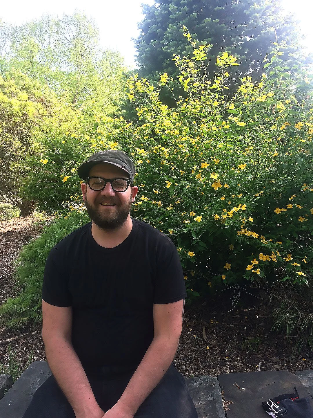 Speiser with glasses, a beard, and a black cap smiling while sitting outdoors in front of green bushes with yellow flowers.