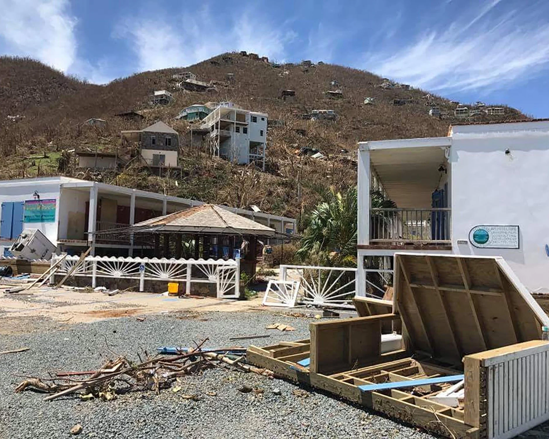 Debris and damaged buildings in Coral Bay after hurricanes, with a hillside of destroyed homes under a partly cloudy sky.