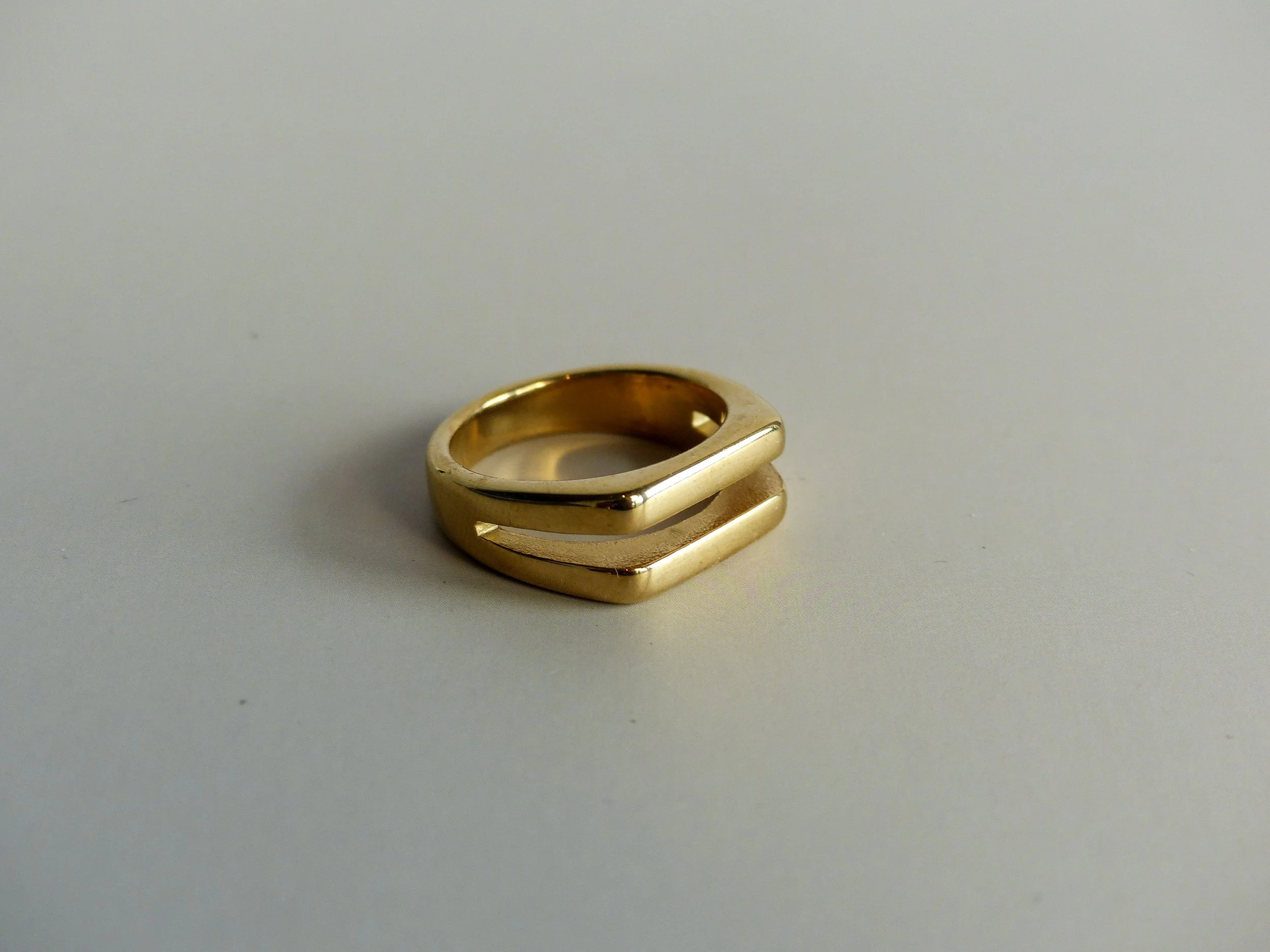 Close-up of a gold ring with a square-shaped top, placed on a plain white surface.