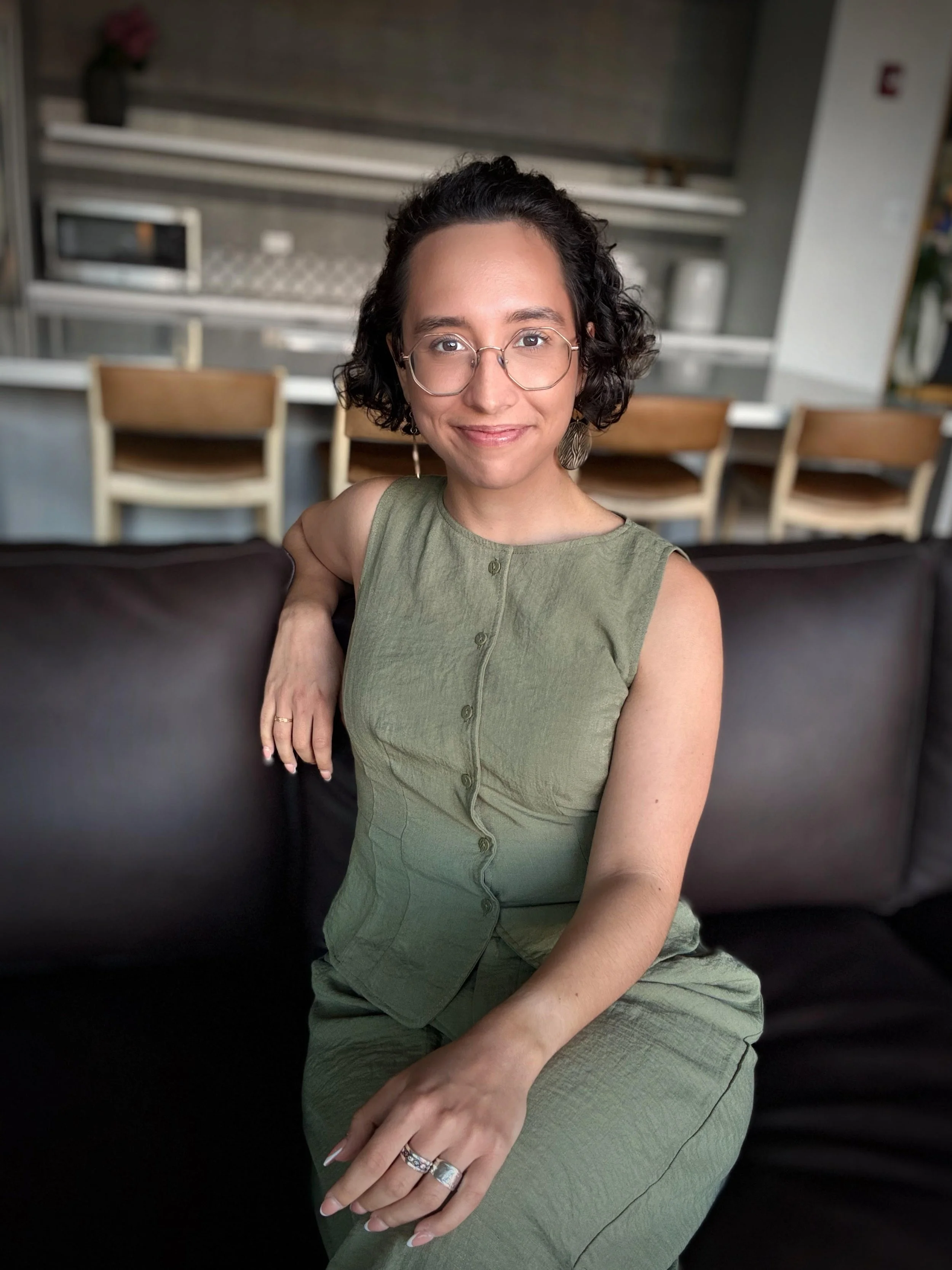 A woman with short curly black hair, wearing glasses and a green sleeveless dress, sitting on a black sofa in a room with shelves and chairs in the background.