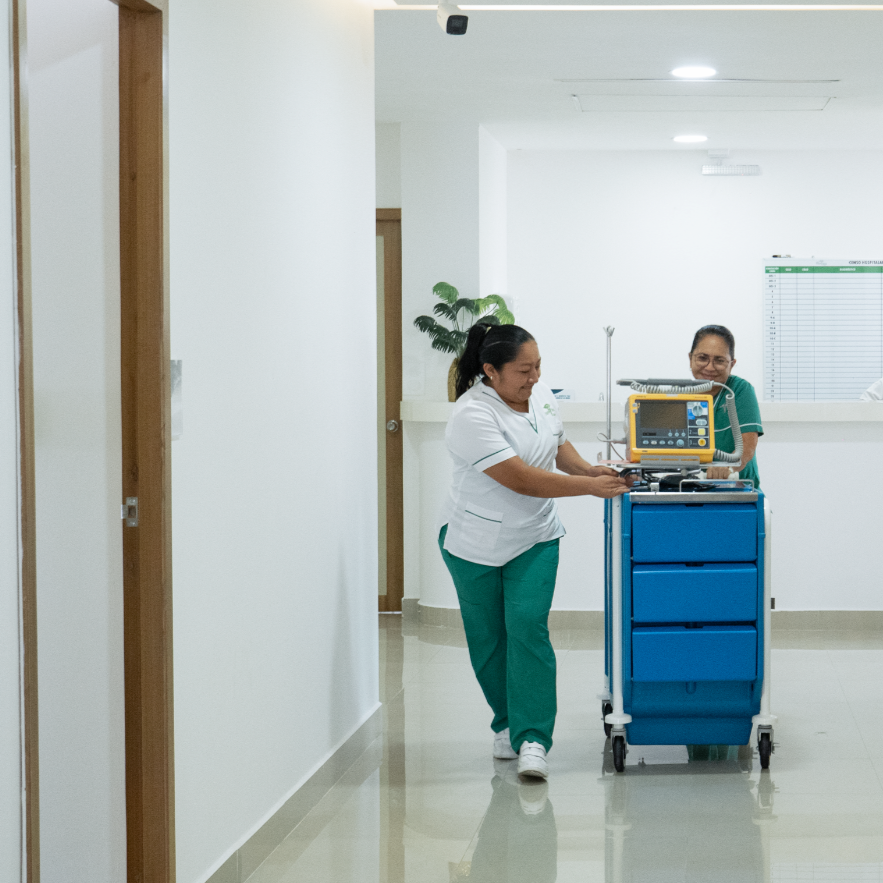 Dos trabajadoras de hospital, una con uniforme blanco y otra con uniforme verde, empujando un carrito con equipo médico en un pasillo de hospital moderno.
