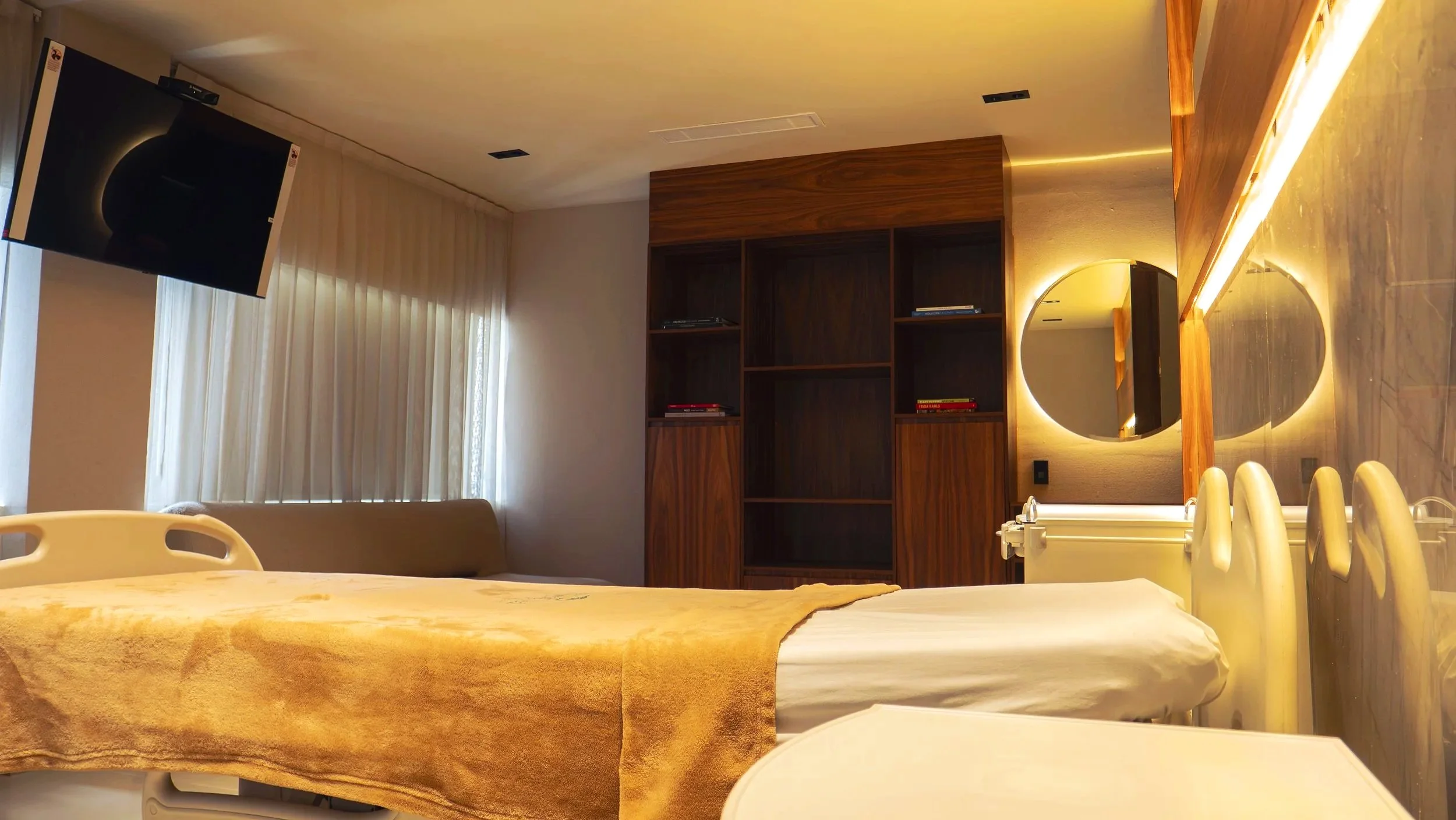 Habitación con cama de hospital, mueble de madera y televisor, con cortinas blancas y luz cálida en la pared con espejo redondo.