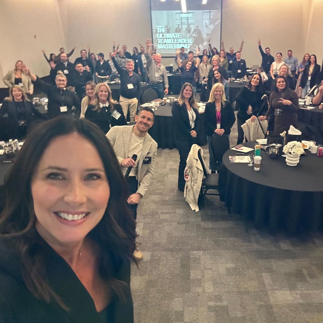 Group of people at a conference in a banquet hall, smiling and waving, with a big screen in the background displaying 'The Ultimate Team Leader Masterclass'.