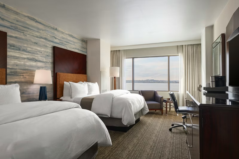 Hotel room with two beds, a large window showing a waterfront view, and standard furniture including a desk, chair, and lamps.