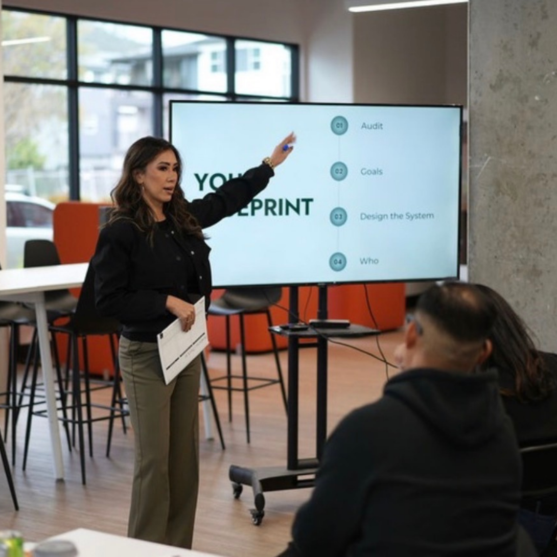 Woman giving a presentation in a meeting room, pointing at a large screen displaying a presentation slide with four bullet points: Audit, Goals, Design the System, and Who.