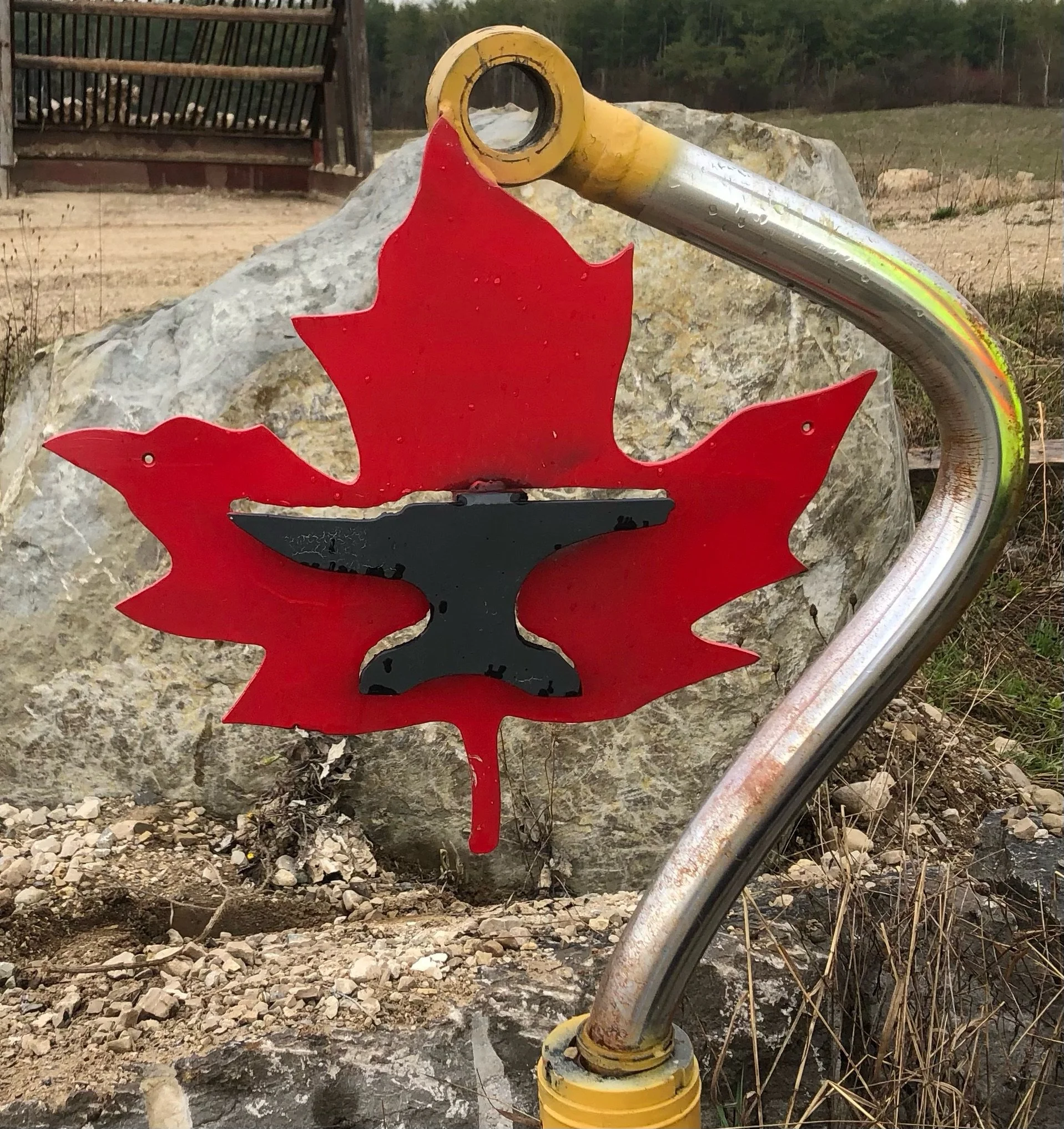 Canadian Iron logo, metal maple leaf with an anvil cutout welded to a bent cylinder rod post.