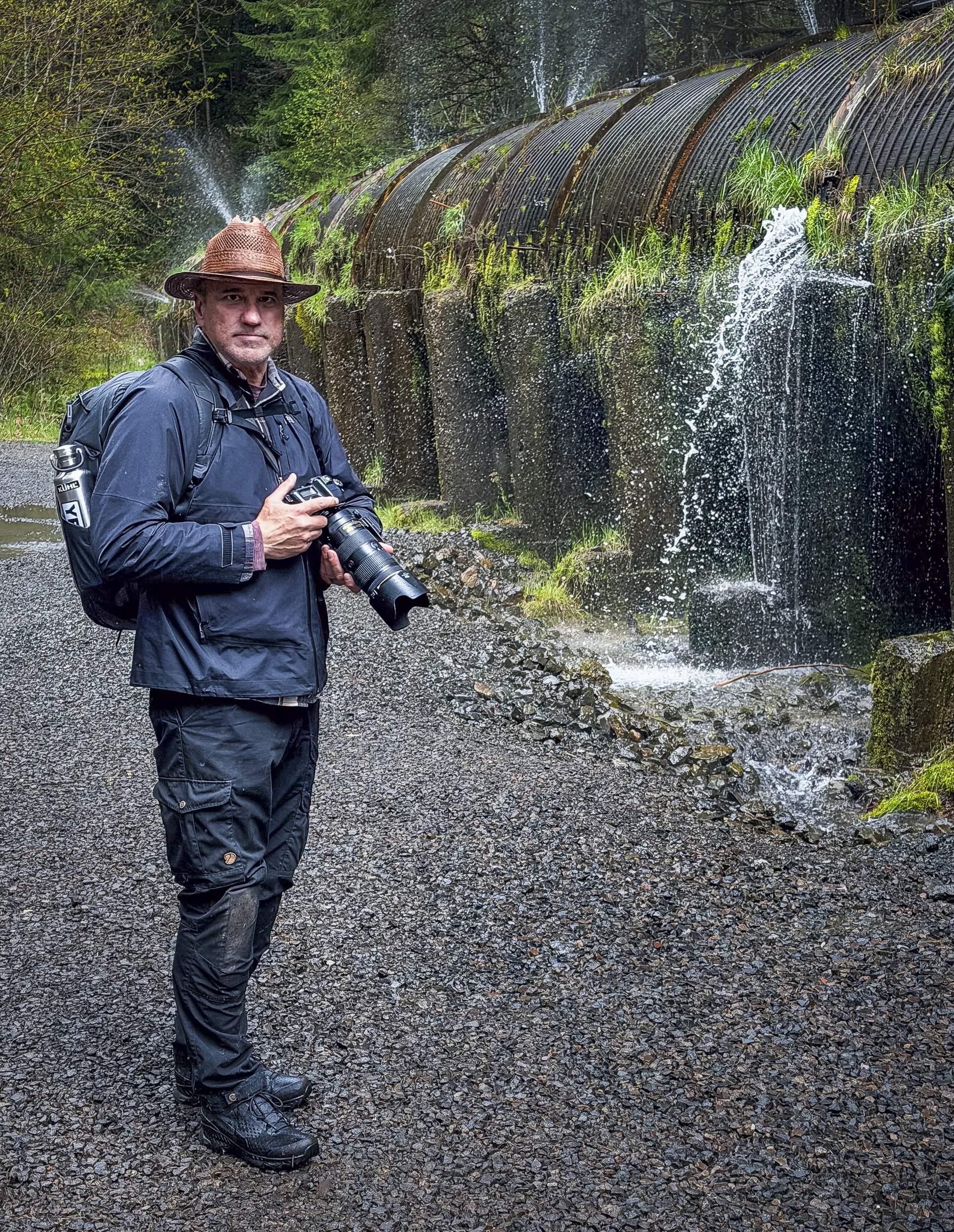 A man wearing a wide-brimmed hat, a black waterproof jacket, and hiking boots stands on a gravel trail holding a camera, with a waterfall flowing down a moss-covered rock face surrounded by lush greenery in the background.