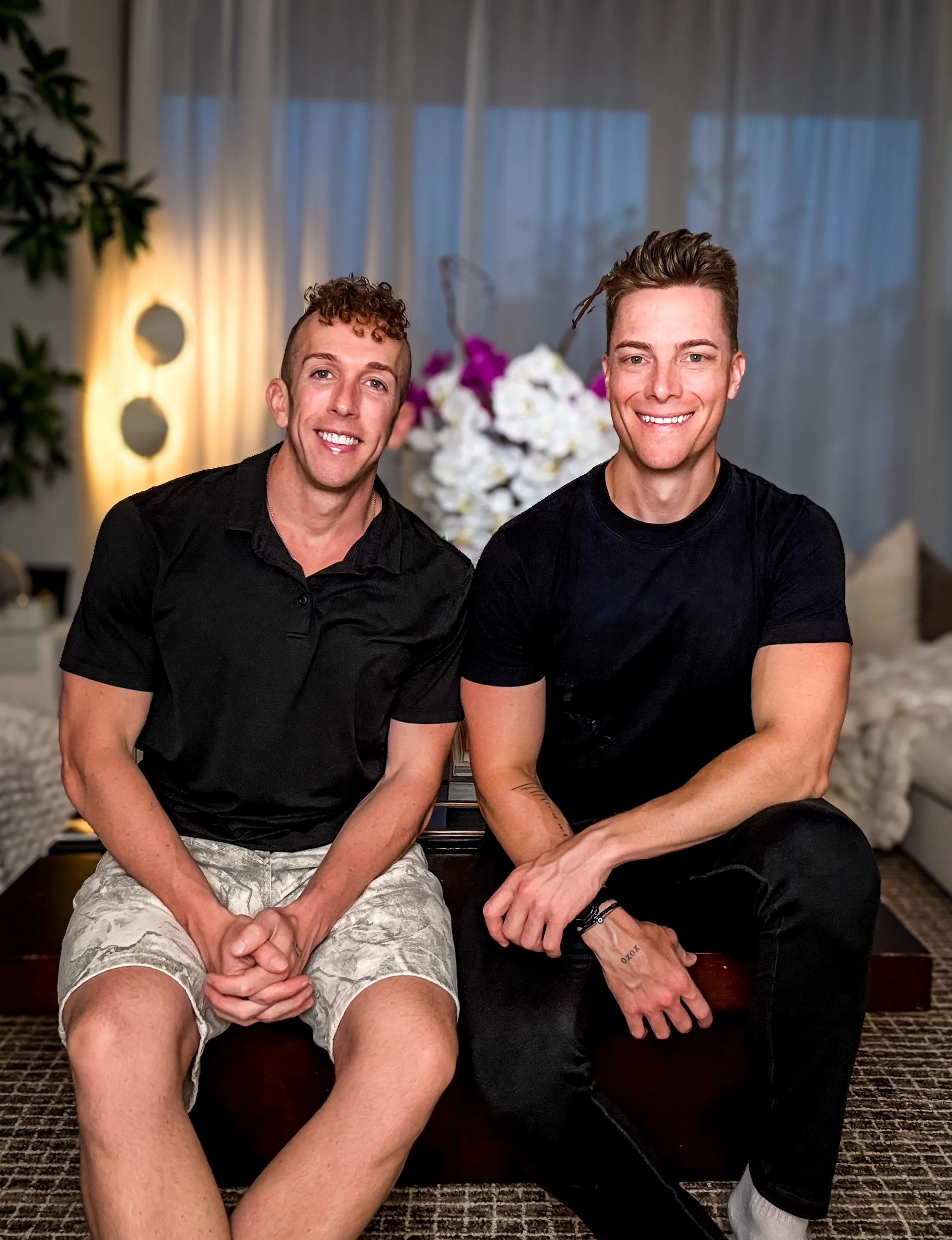 Two young men sitting close together indoors, smiling at the camera, with floral decor and a window with curtains in the background.