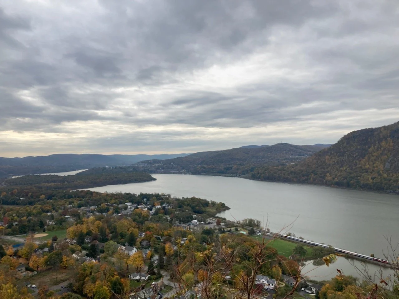 A scenic view of a river winding through a town with hills covered in fall foliage, under a cloudy sky.