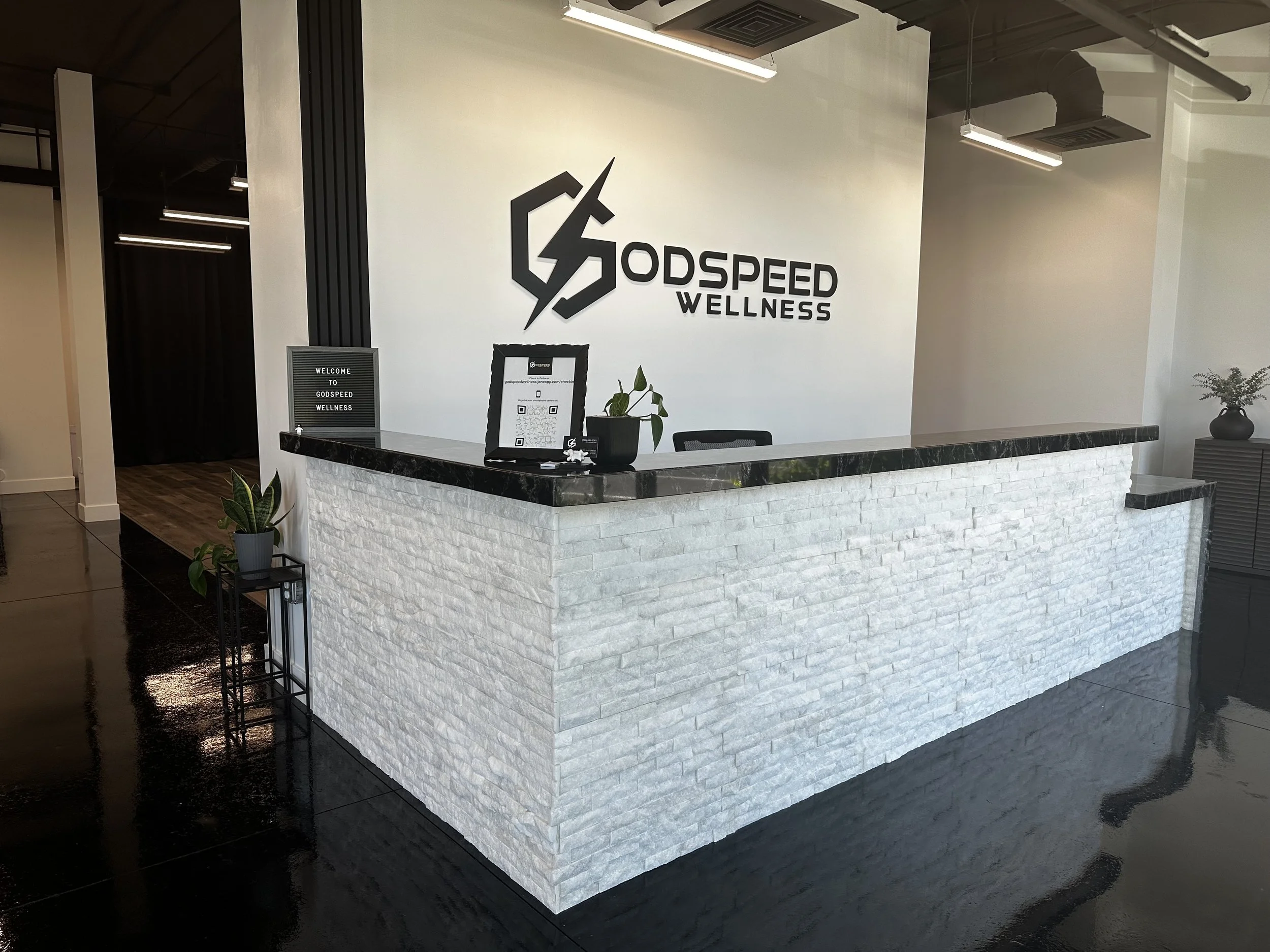 Reception desk with a black marble top, white brick base, and a sign on the wall behind that reads 'GODSPEED WELLNESS' with a logo. There are potted plants on the desk and a black chair behind it, in a modern wellness or fitness center.