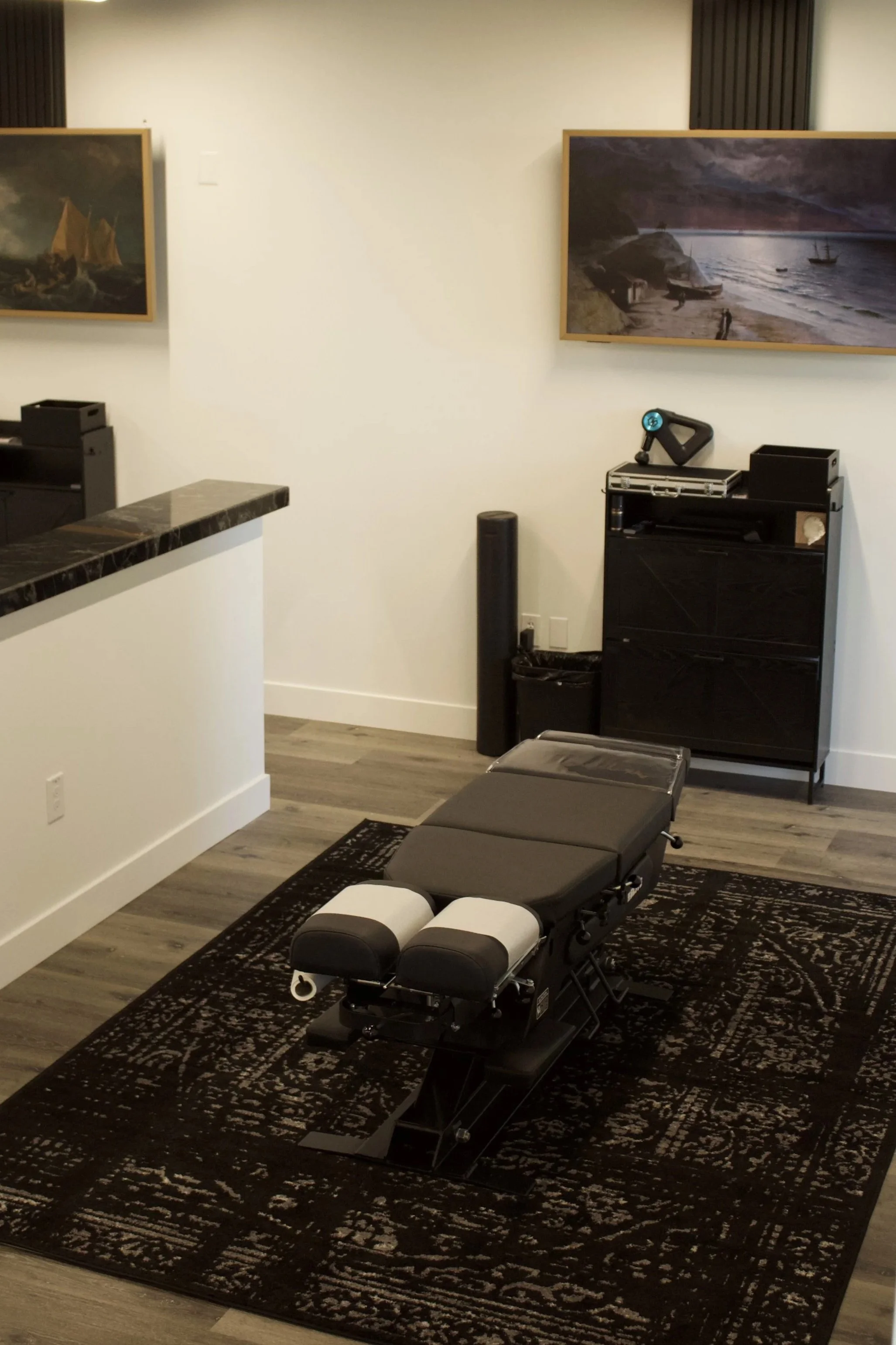 Interior view of a massage or therapy room with a massage table, black carpet, framed paintings of seascapes, and white walls.