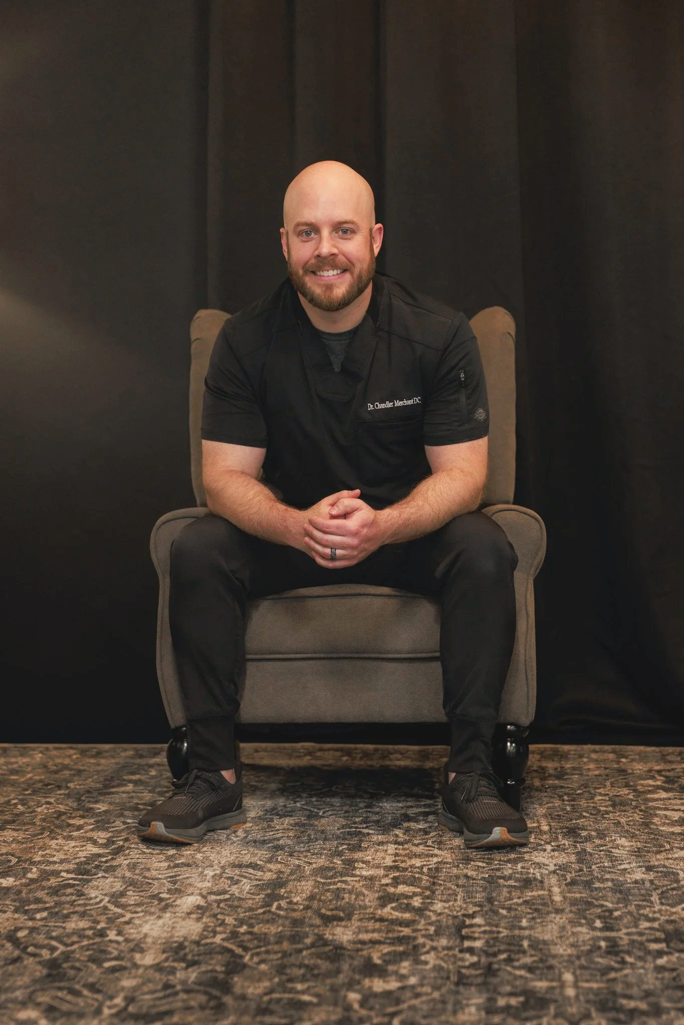 A man with a beard, wearing a black shirt and black pants, sitting on a beige armchair in front of a dark background, smiling at the camera.