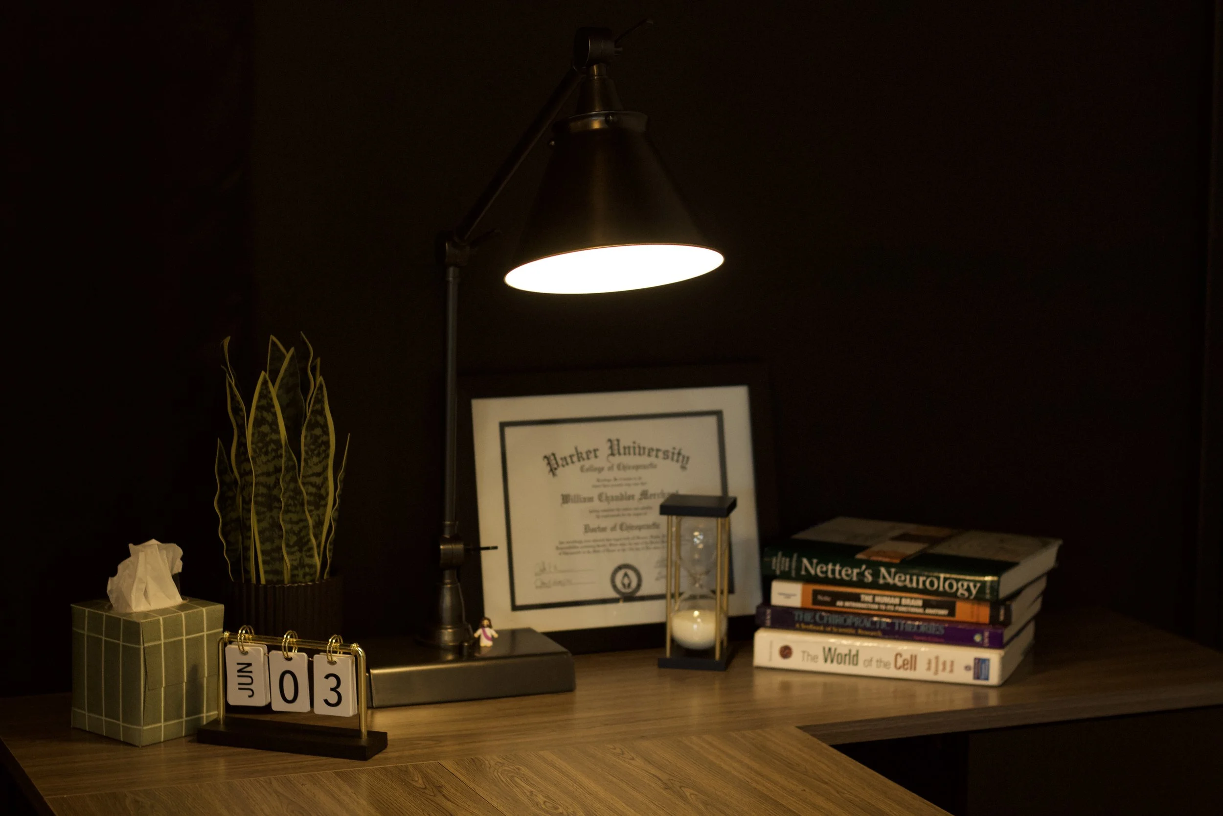 A desk displaying various books, a framed certificate, a small clock, a decorative plant, a tissue box, a calendar, and a lampshade.