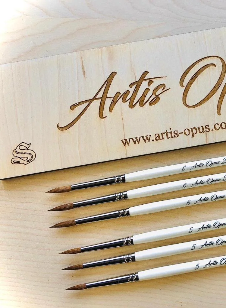 Six paintbrushes with brown bristles and white handles, labeled with sizes 5 and 6, are laid out on a light wooden surface in front of a wooden box with the brand name Ignite, website, and logo.