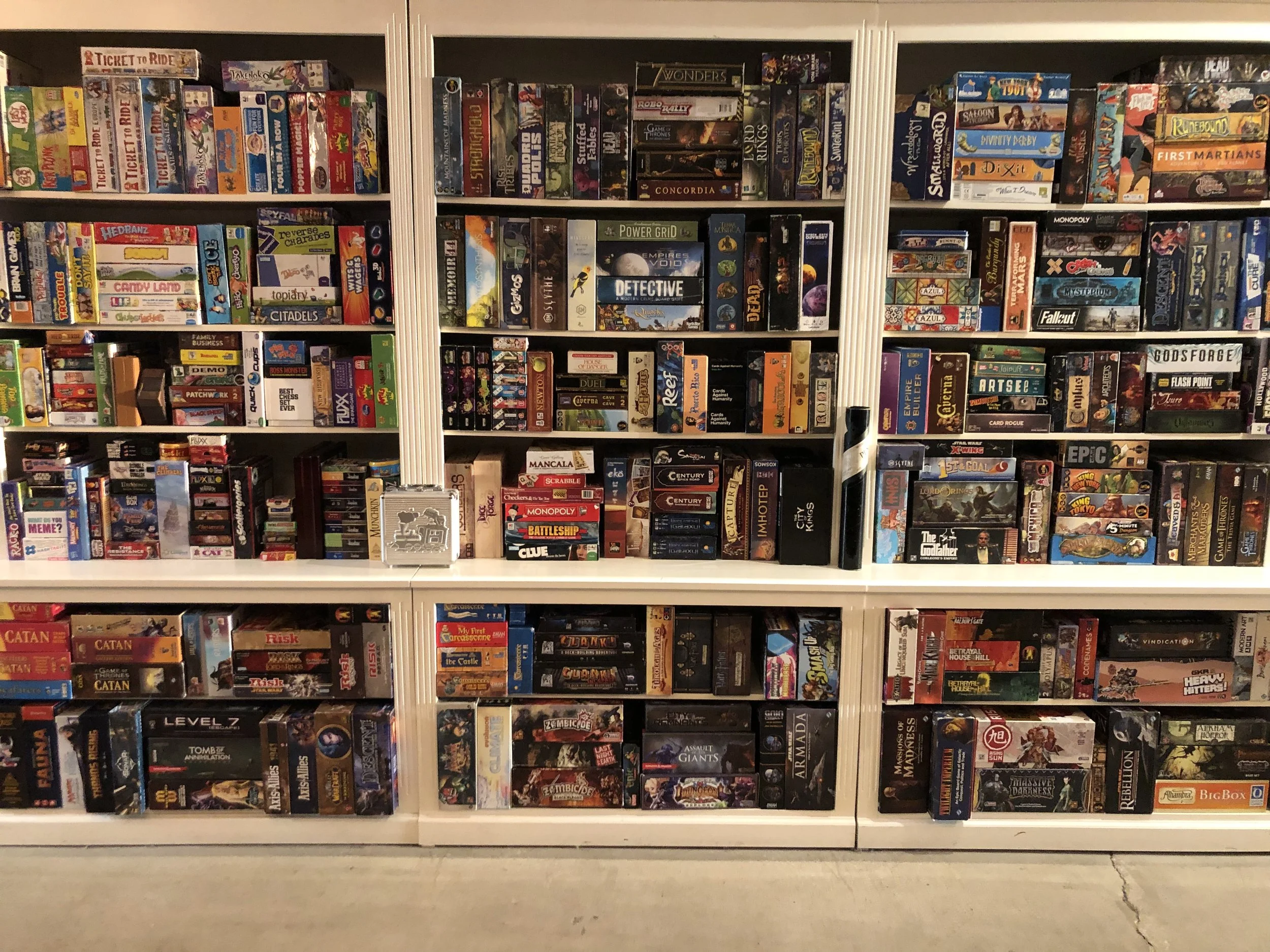 A white bookshelf filled with tabletop and card games, organized by genre and size, in a well-lit room.