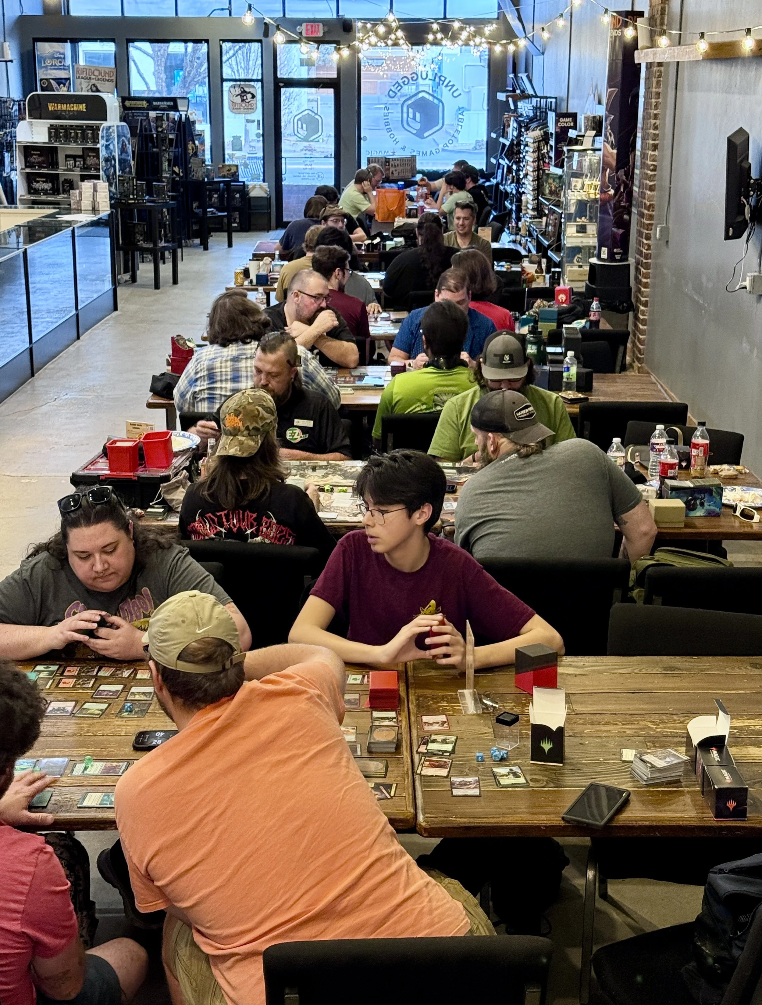 People playing a tabletop card game at a gaming store, with shelves of board games and comic books in the background, and large windows showing a snowy outdoors.