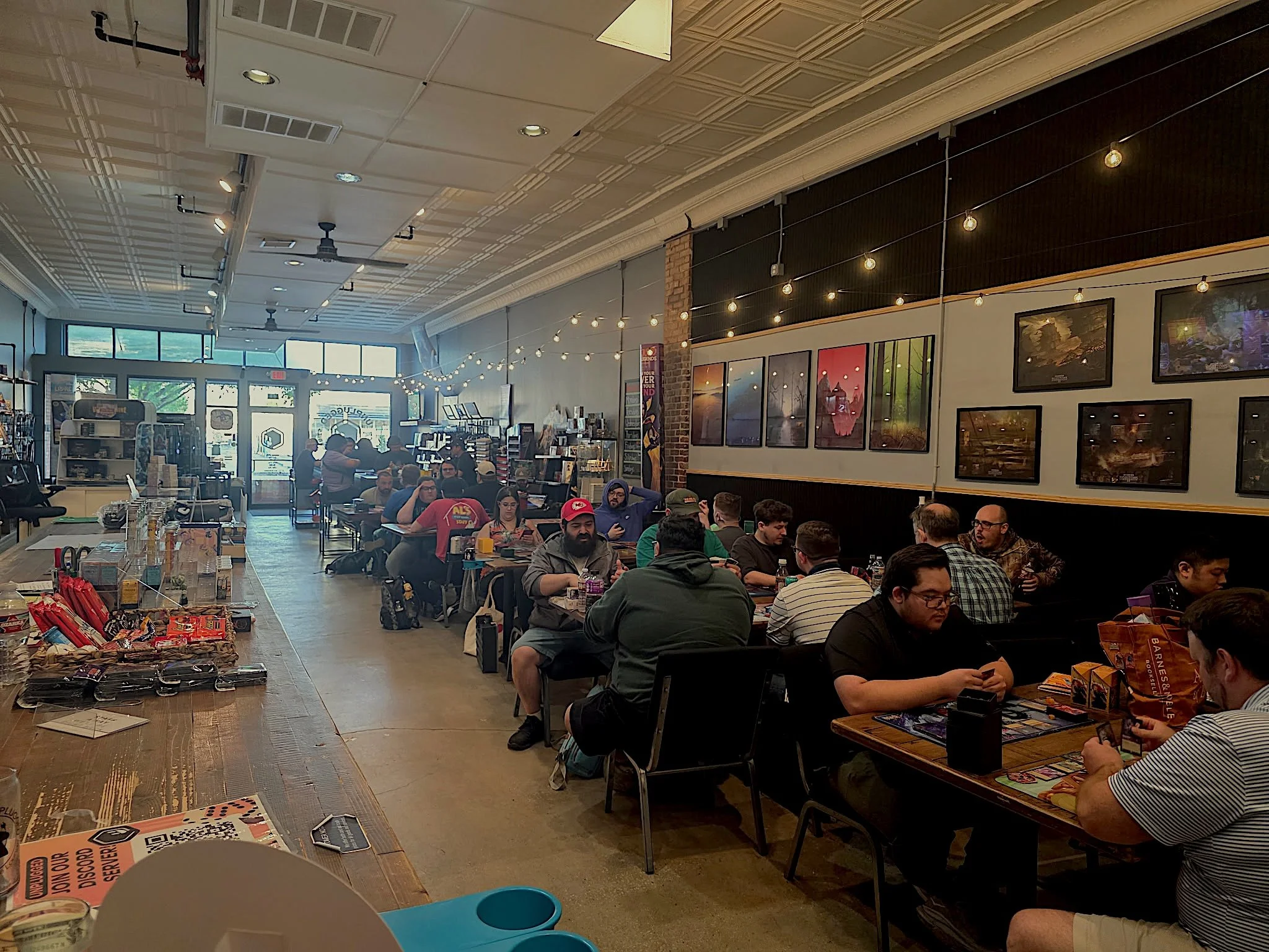A crowded room with many people sitting at tables, playing card games, inside a store or cafe with art on the walls and string lights hanging from the ceiling.