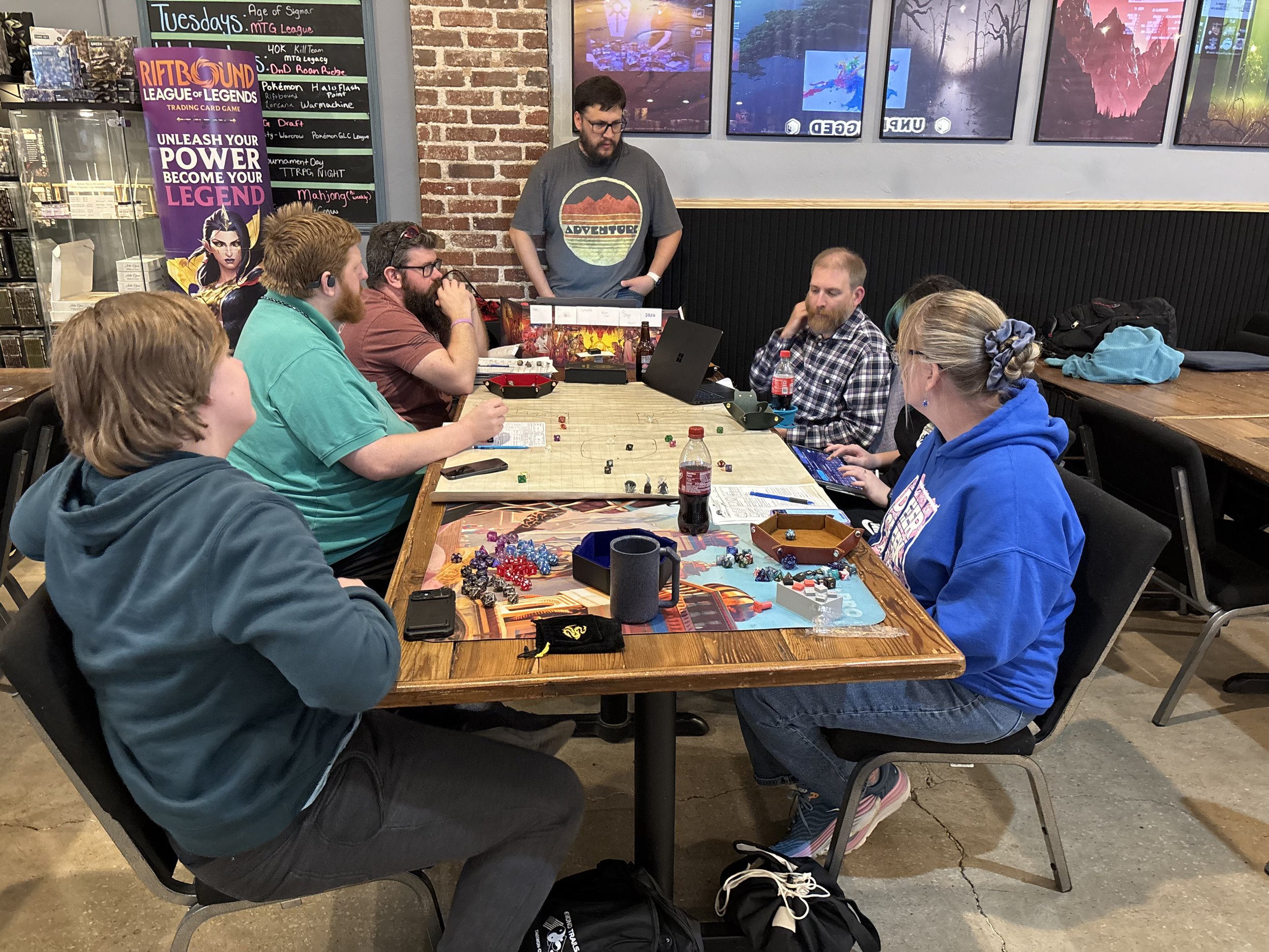 A group of seven people sit around a table playing tabletop games at a restaurant or cafe. There are dice, game boards, and drinks on the table. A man, standing, wears a t-shirt with an adventure graphic, and several other individuals are looking at their game materials or at the center of the table. There are large windows, a brick wall, and a colorful poster for a trading card game in the background.